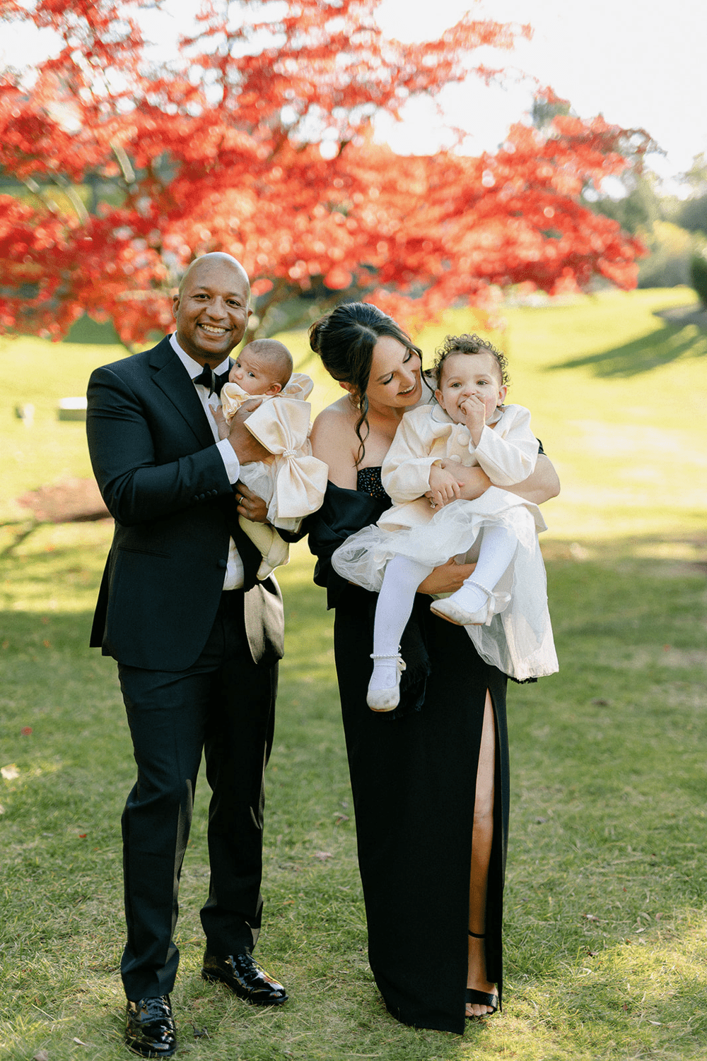 Family of four in formal attire standing outdoors with red autumn leaves in the background.
