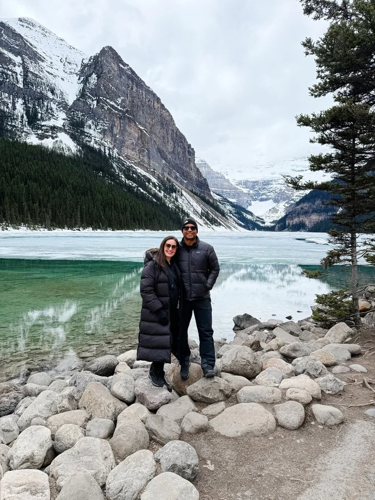 A couple standing on rocks by a lake surrounded by snow-covered mountains and trees.