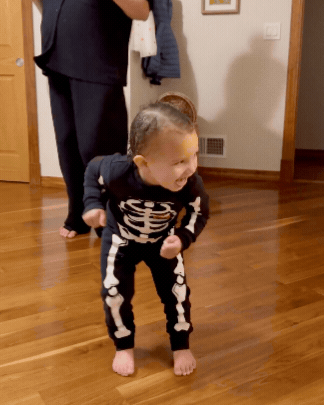 A young child in a skeleton costume standing on a hardwood floor, smiling and in a playful pose indoors, with an adult and another child partially visible in the background.
