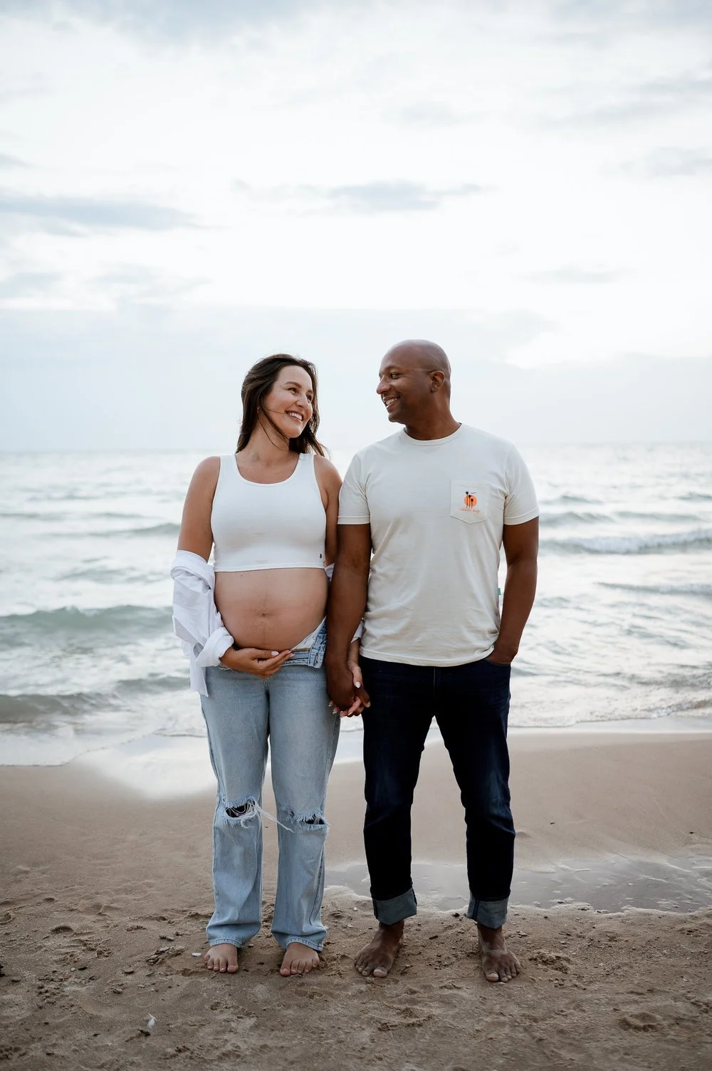 A pregnant woman and her partner standing barefoot on the beach, holding hands, smiling, with the ocean and cloudy sky in the background.