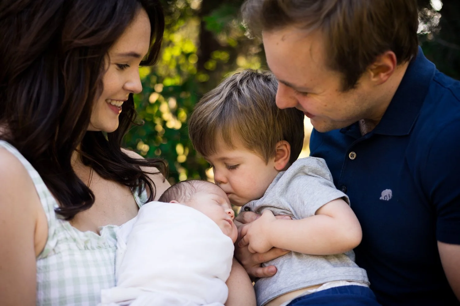 A family of four, including a mother, father, an older boy, and a newborn baby, enjoying a tender moment outdoors with greenery in the background. The mother is holding the newborn, and the older boy is gently touching their foreheads as the father looks on with a smile.