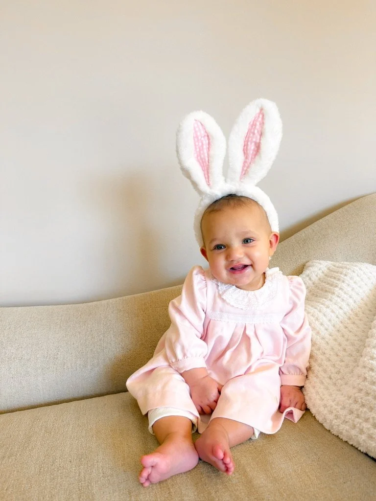 A baby girl wearing bunny ears headband and pink dress, sitting on a beige couch, smiling.