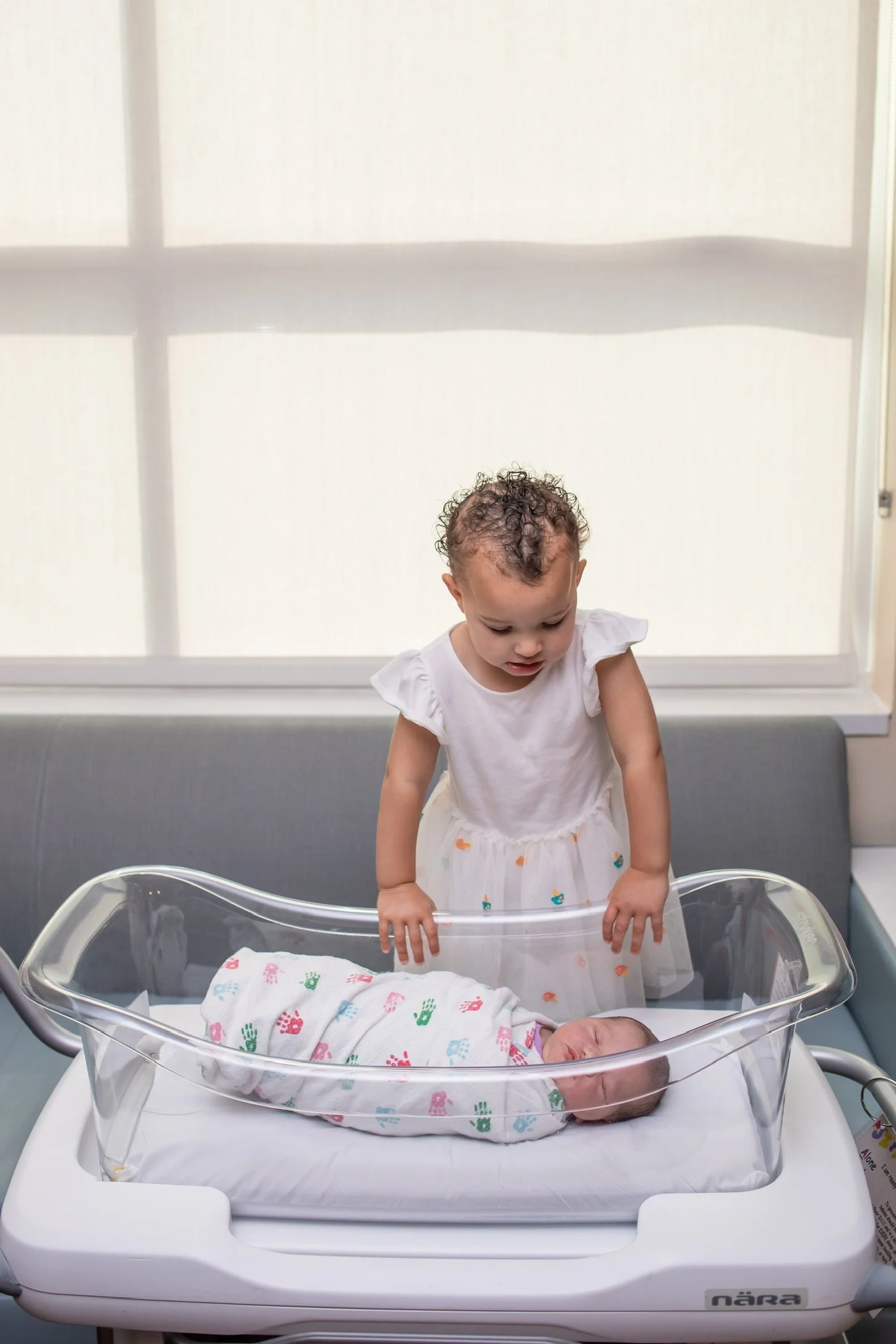 A young girl with curly hair and a white dress stands beside a hospital bassinet, looking down at a newborn baby wrapped in a colorful blanket inside the bassinet.