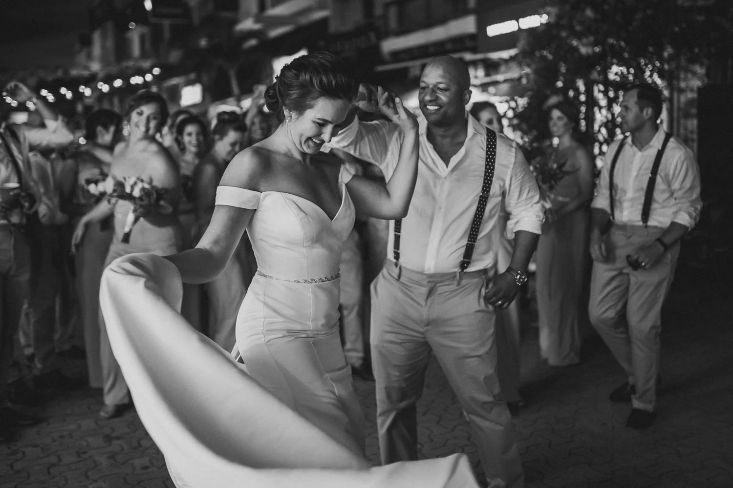 A woman in a white off-shoulder wedding dress is dancing with a man in a white shirt with suspenders. They are smiling and laughing, surrounded by other people at a celebration or wedding reception, captured in black and white.