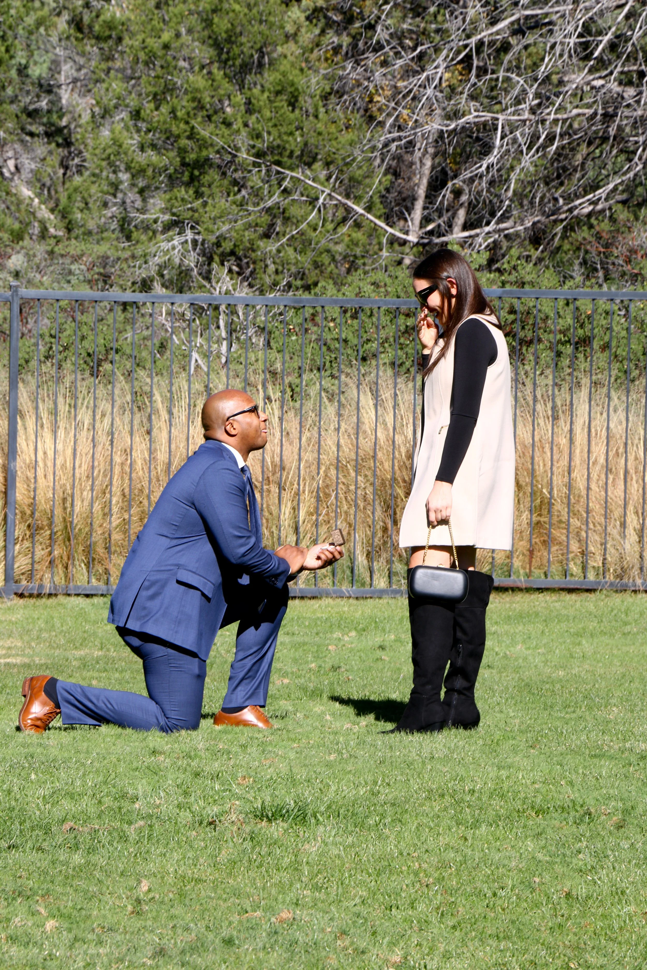 A man kneeling on one knee proposing marriage to a woman outdoors, with a fence and trees in the background.