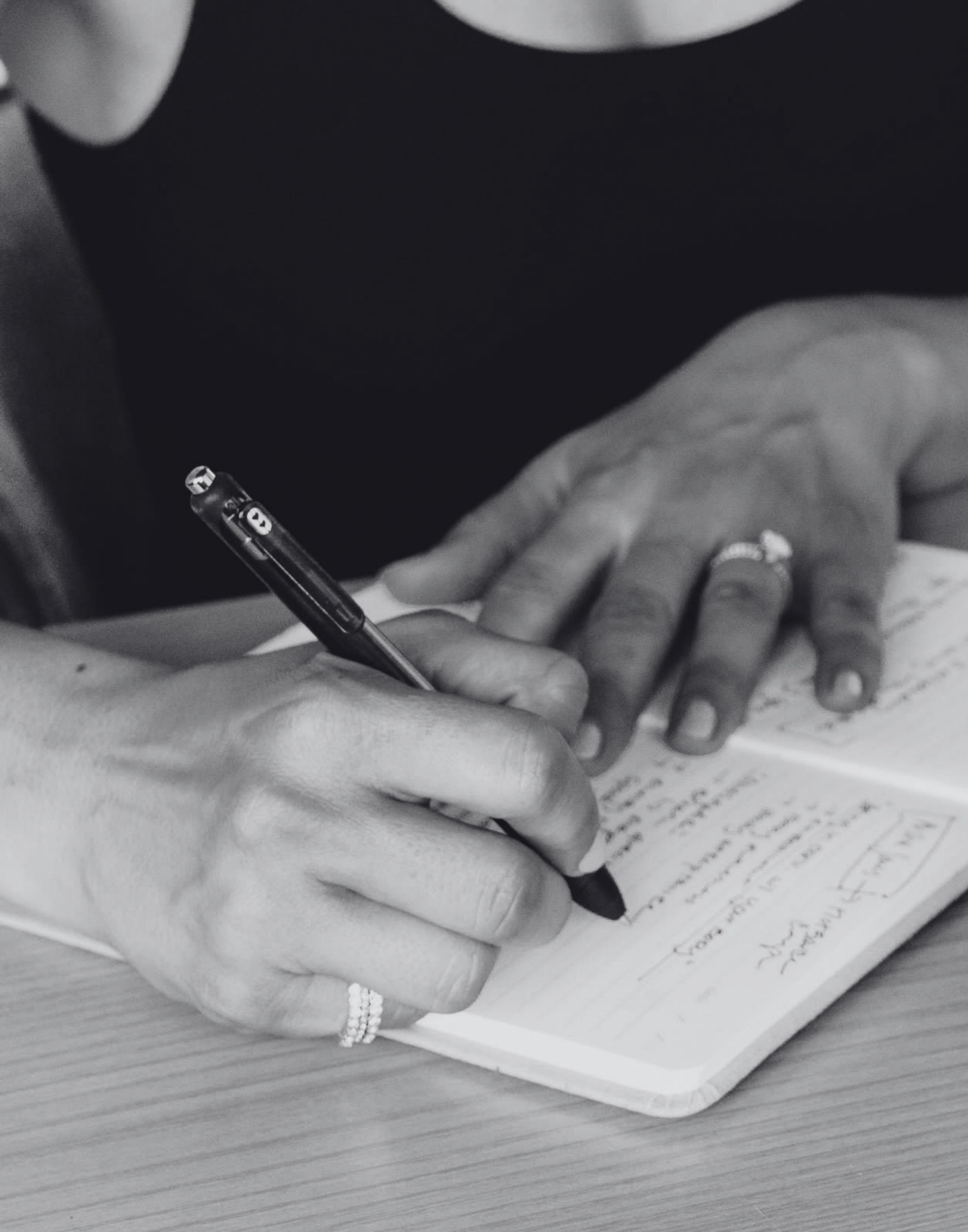 Close-up of a hand holding a pen writing on a piece of paper, with the edge of a notebook visible on a wooden surface.