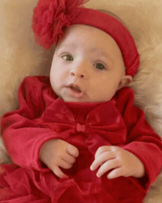 A baby wearing a red headband with a large bow and a red dress, lying on a soft surface.