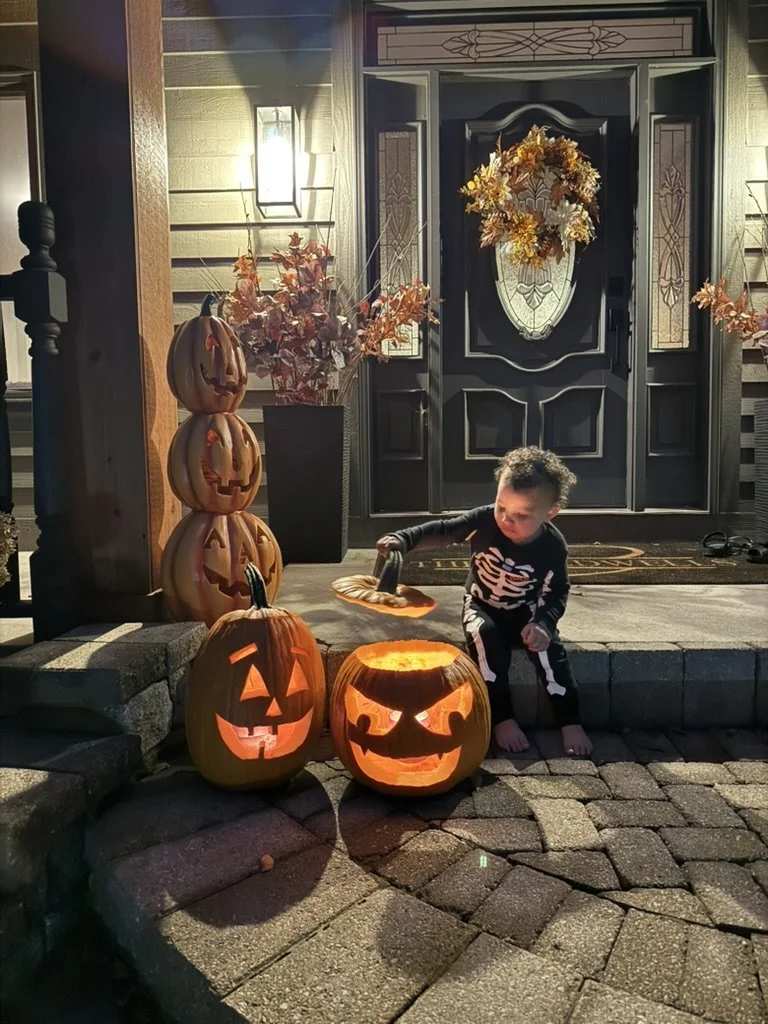 A young boy dressed in a skeleton costume sits on a brick step outside a house, holding a small Halloween-themed plate. Two lit jack-o'-lantern pumpkins with carved faces are in front of him. To his left are stacked pumpkins with carved faces. The front door is decorated with an autumn wreath, and there are autumn leaves in a tall vase next to a wall-mounted lantern light.