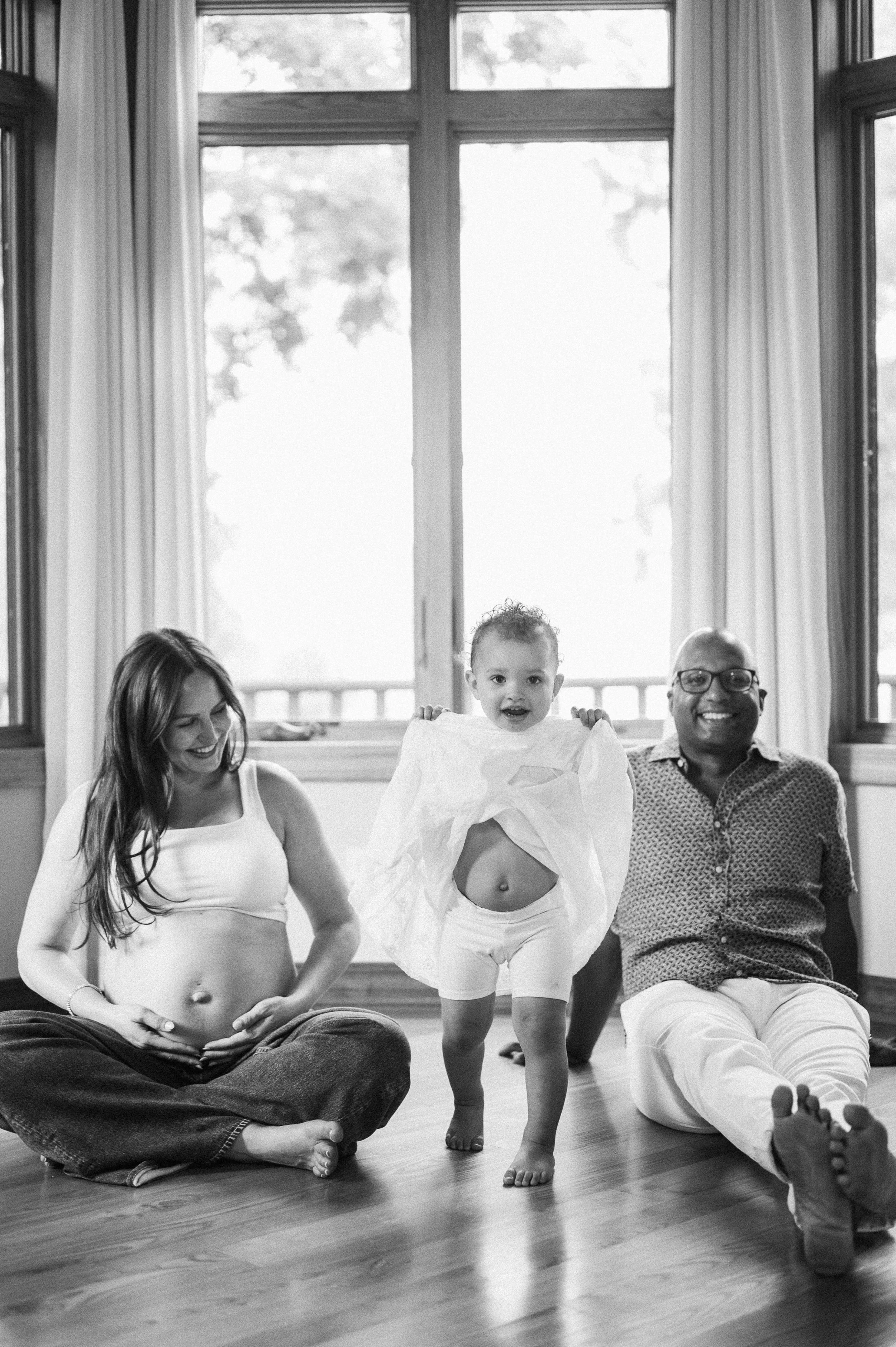 A family of four sitting on the floor in front of a large window, with the mother appearing pregnant, the father smiling, and a young girl standing in the middle lifting a piece of cloth, inside a bright room with curtains.