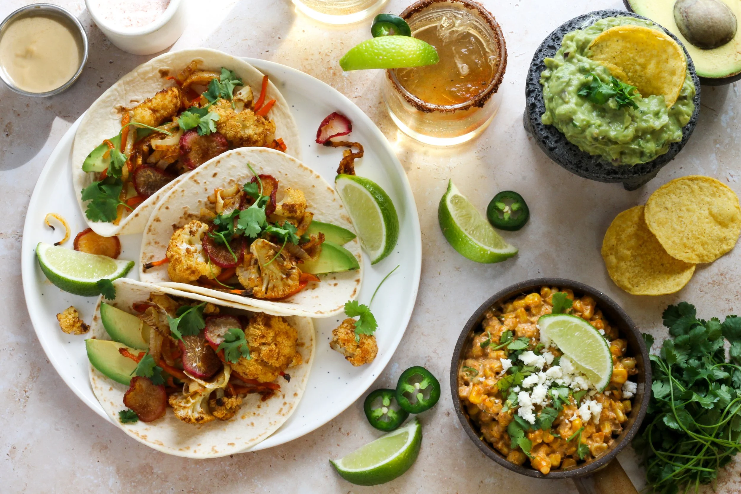 Plate of three tacos filled with fried cauliflower, vegetables, and cilantro, accompanied by lime wedges. Surrounding dishes include guacamole, a drink with a lime wedge and salted rim, yellow corn chips, and a bowl of corn salad with lime, cheese, and herbs. Lime slices and jalapeno rings are scattered around the table.