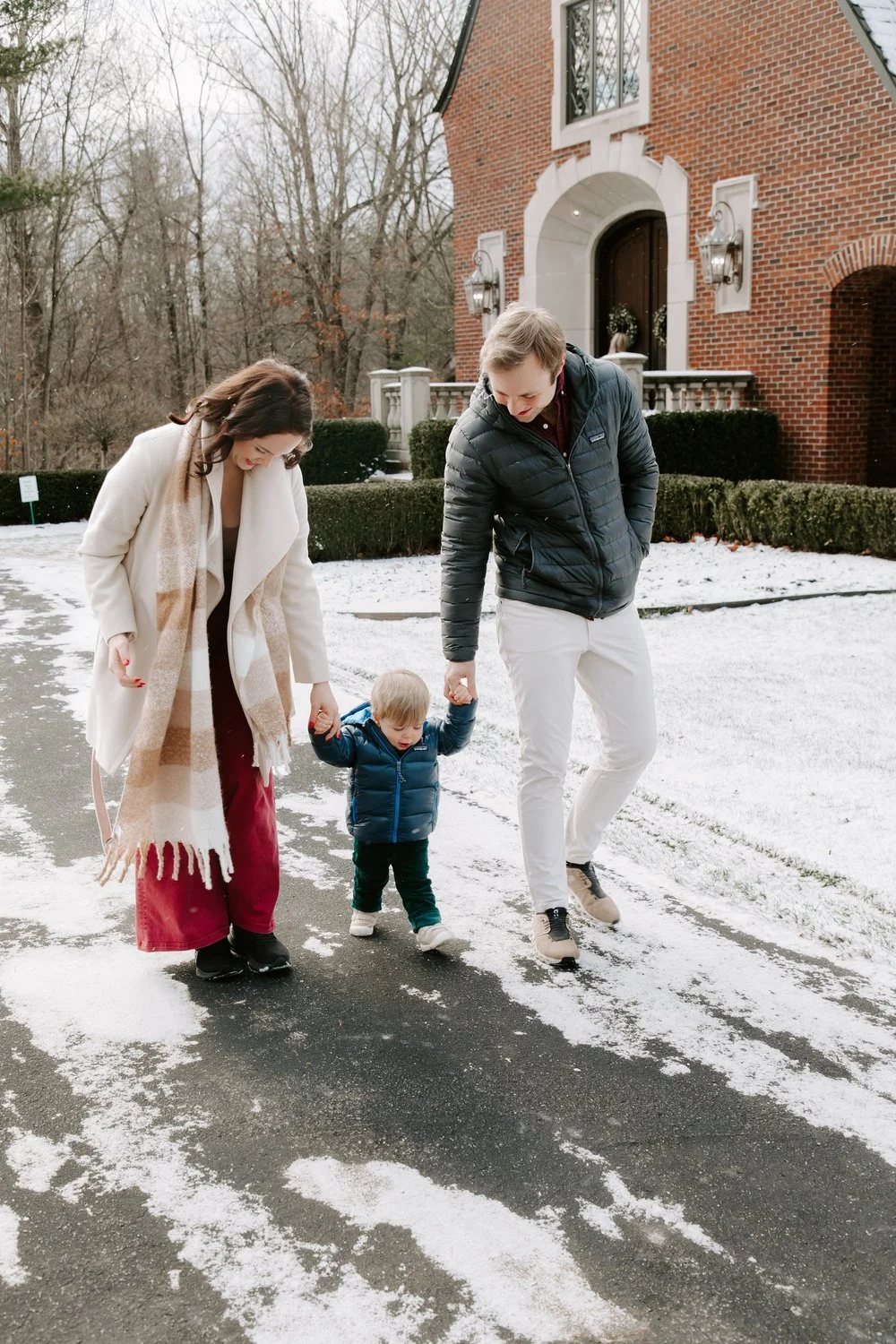 Family of three, mother, father, and young child, walking on a snow-covered path outside a large brick house during winter.