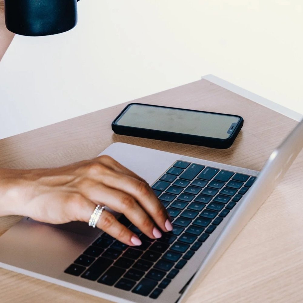 Person typing on a MacBook keyboard with a smartphone and a coffee mug on a wooden desk.