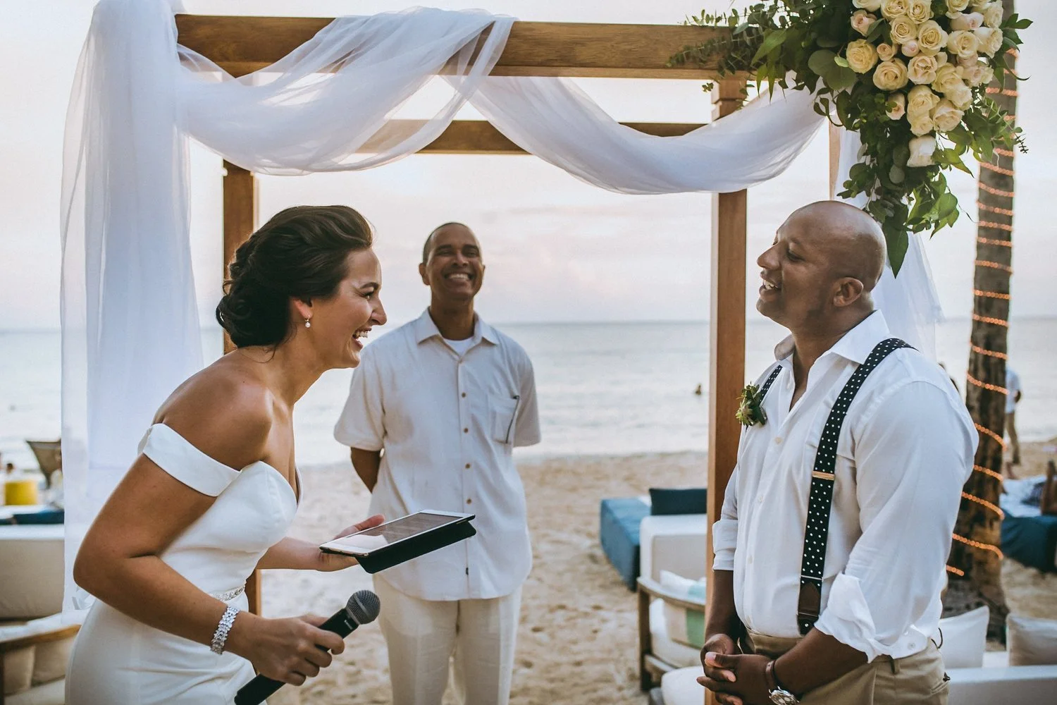 A bride and groom are smiling and exchanging vows during a beach wedding ceremony, with a woman officiant holding a microphone and reading from a tablet, and a man standing behind them. The ceremony is outdoors near the ocean, decorated with a wooden arch draped with white fabric and a floral arrangement.