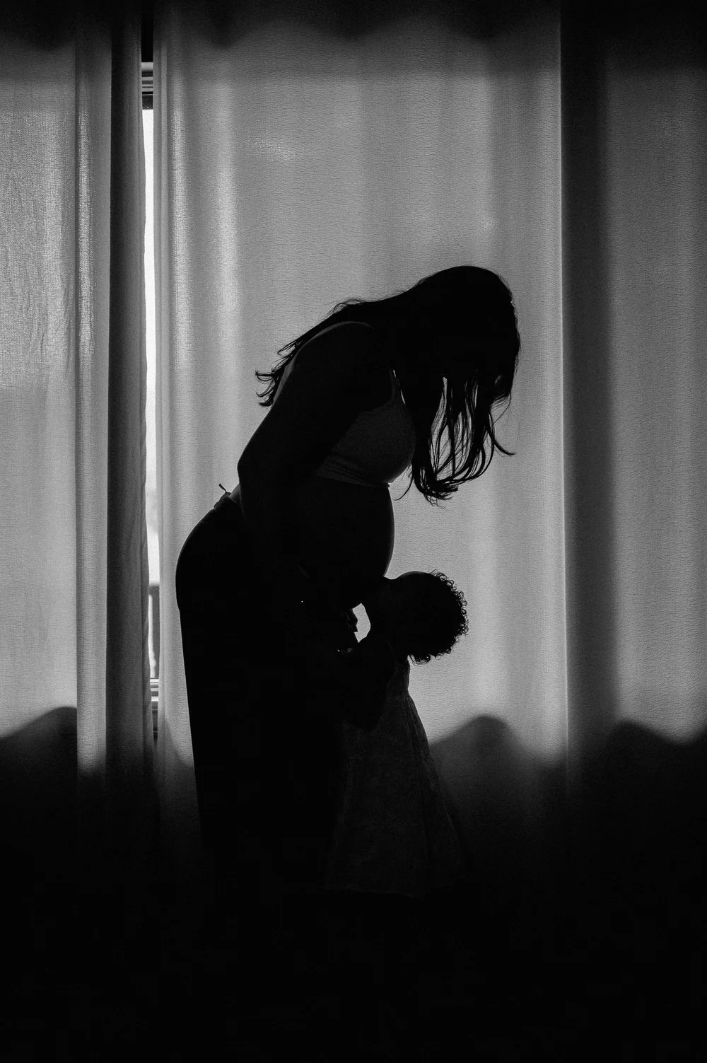 Silhouette of a woman gently holding a child, standing in front of a window with curtains, in black and white.