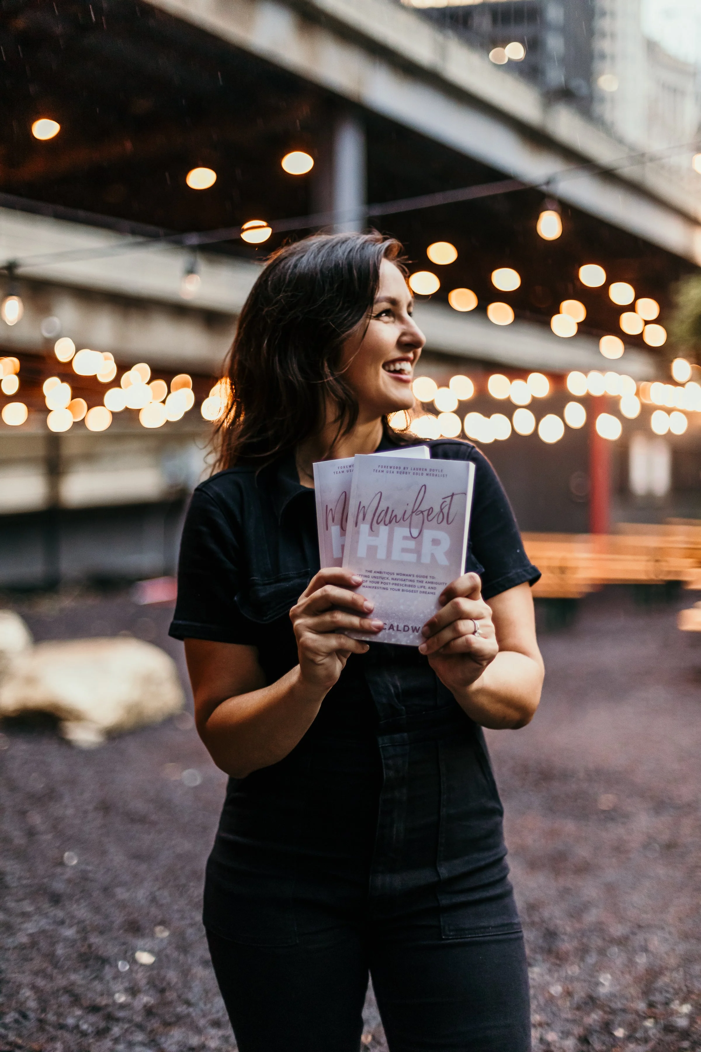 A woman with dark hair smiling, holding a book titled 'Manifest Her' outdoors at dusk, with strings of warm lights overhead and a blurred cityscape in the background.
