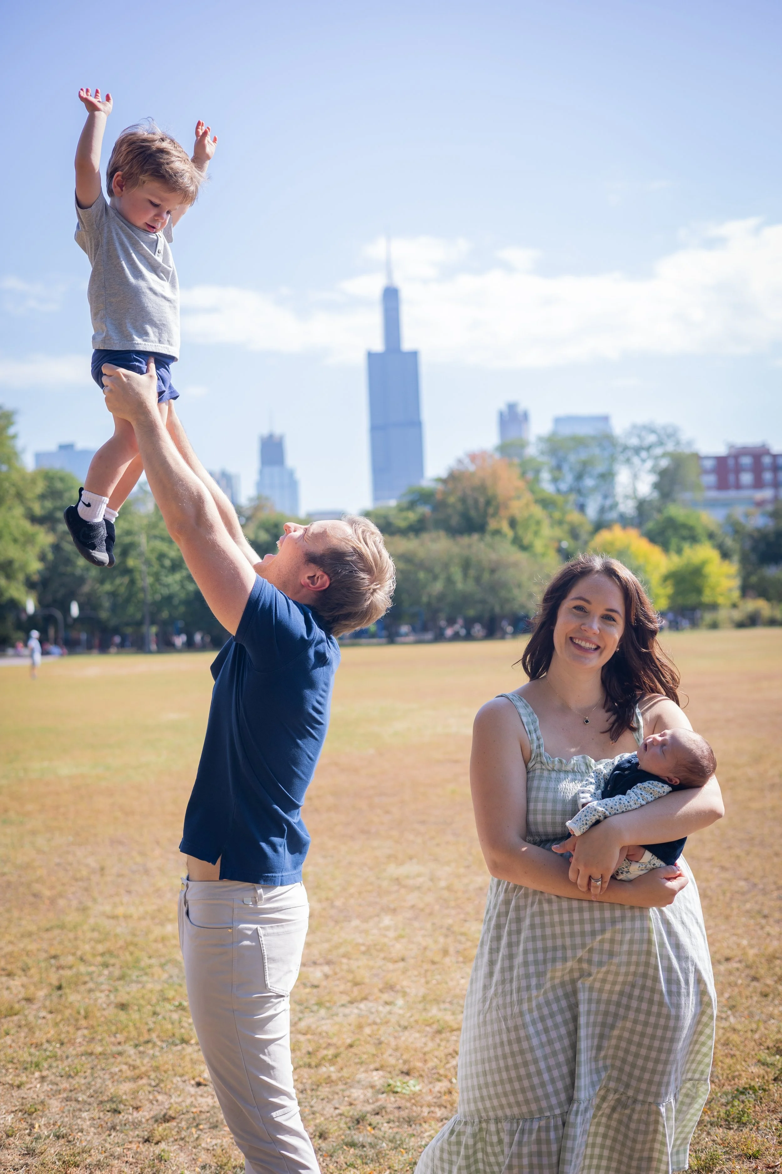 Happy family enjoying a day in the park with a father lifting his son high in the air while a mother holds a baby, city skyline in the background, sunny weather, green trees, and a grassy field.