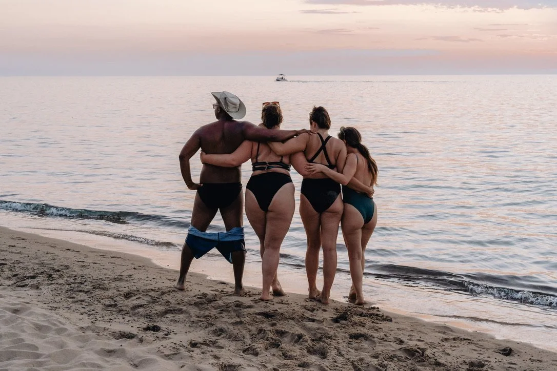 Four friends embracing on the beach during sunset, standing by the water with a boat in the distance.