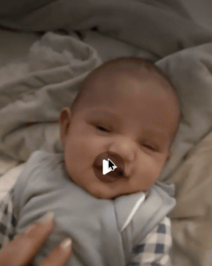 A baby lying in bed with a pacifier in their mouth, looking towards the camera.
