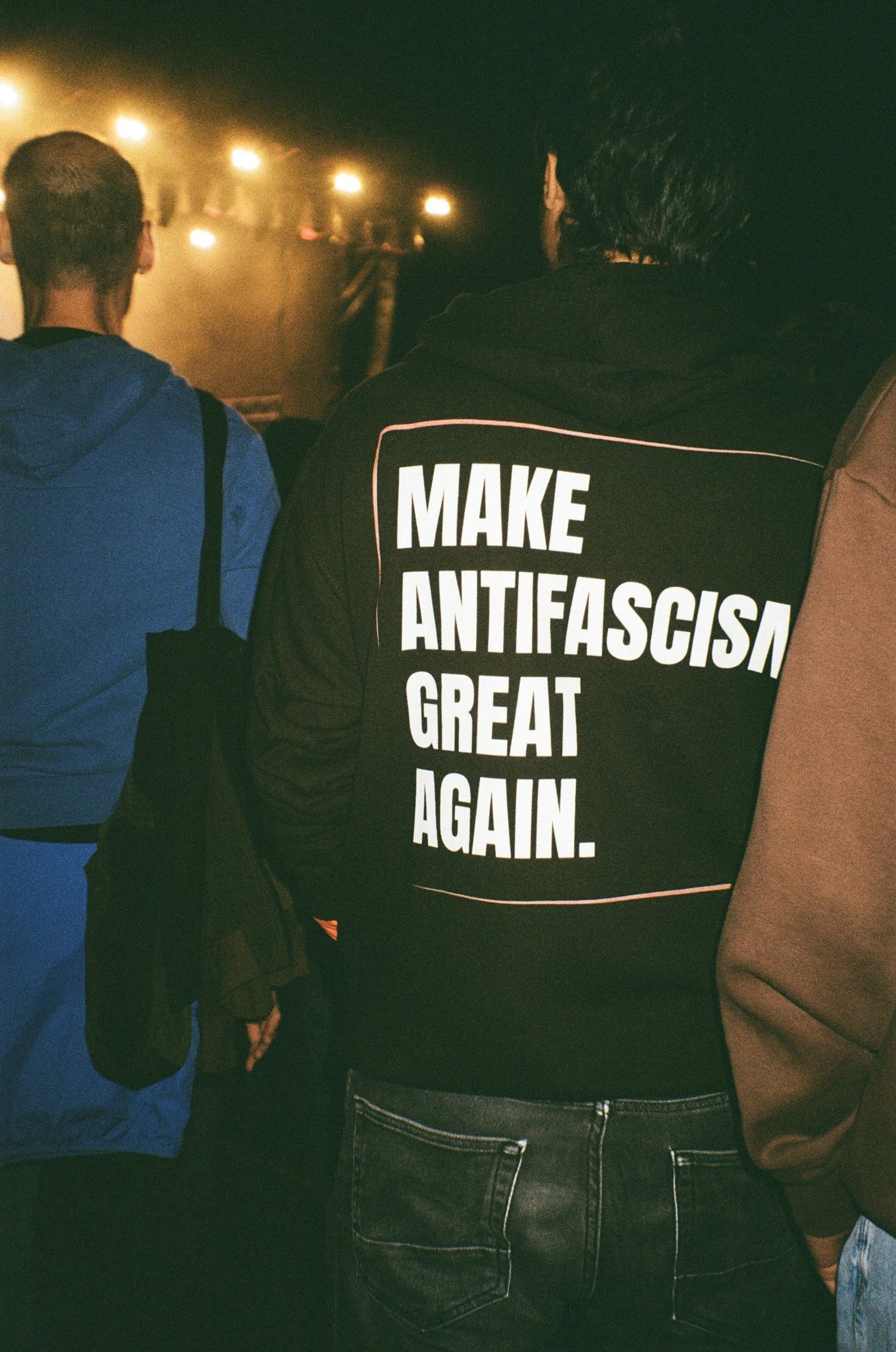 the back of three people wearing sweatshirts close up, the middle shirt has script 'make antifascism great again.'