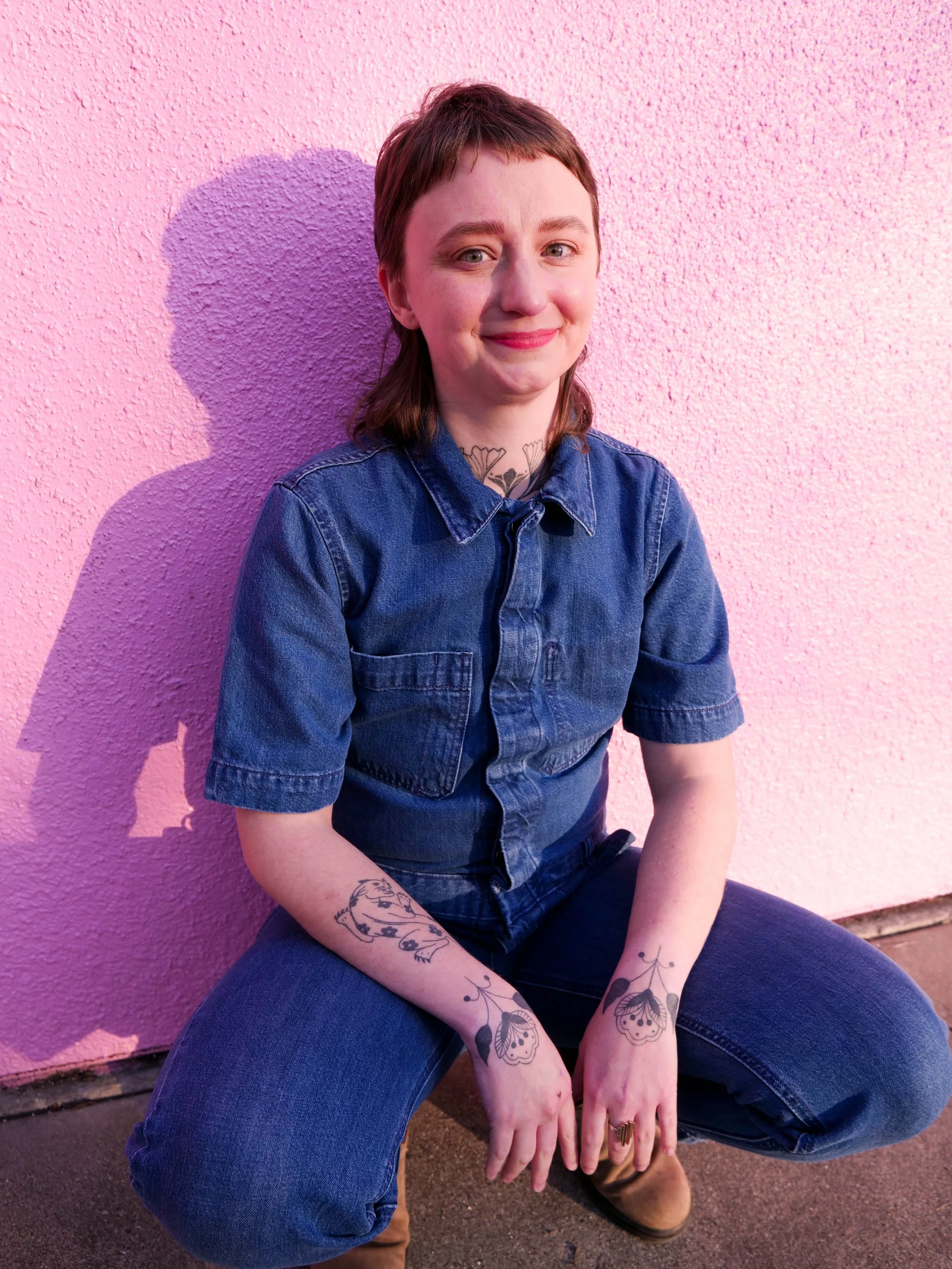 white, non-binary person, with brown hair cut into a mullet, wearing denim jumpsuit and squatting in front of a bubblegum pink wall. sun on their face and shadow to the left of the frame.