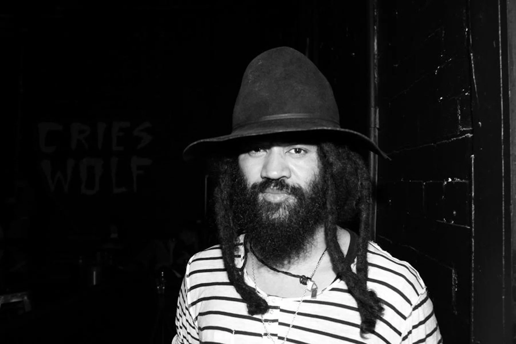A black-and-white photo of a man with a large beard, wearing a wide-brimmed hat, striped shirt, and dreadlocks, standing in front of a dark background with some visible wooden panels and a partially visible sign that reads 'CRIES WOLF'.