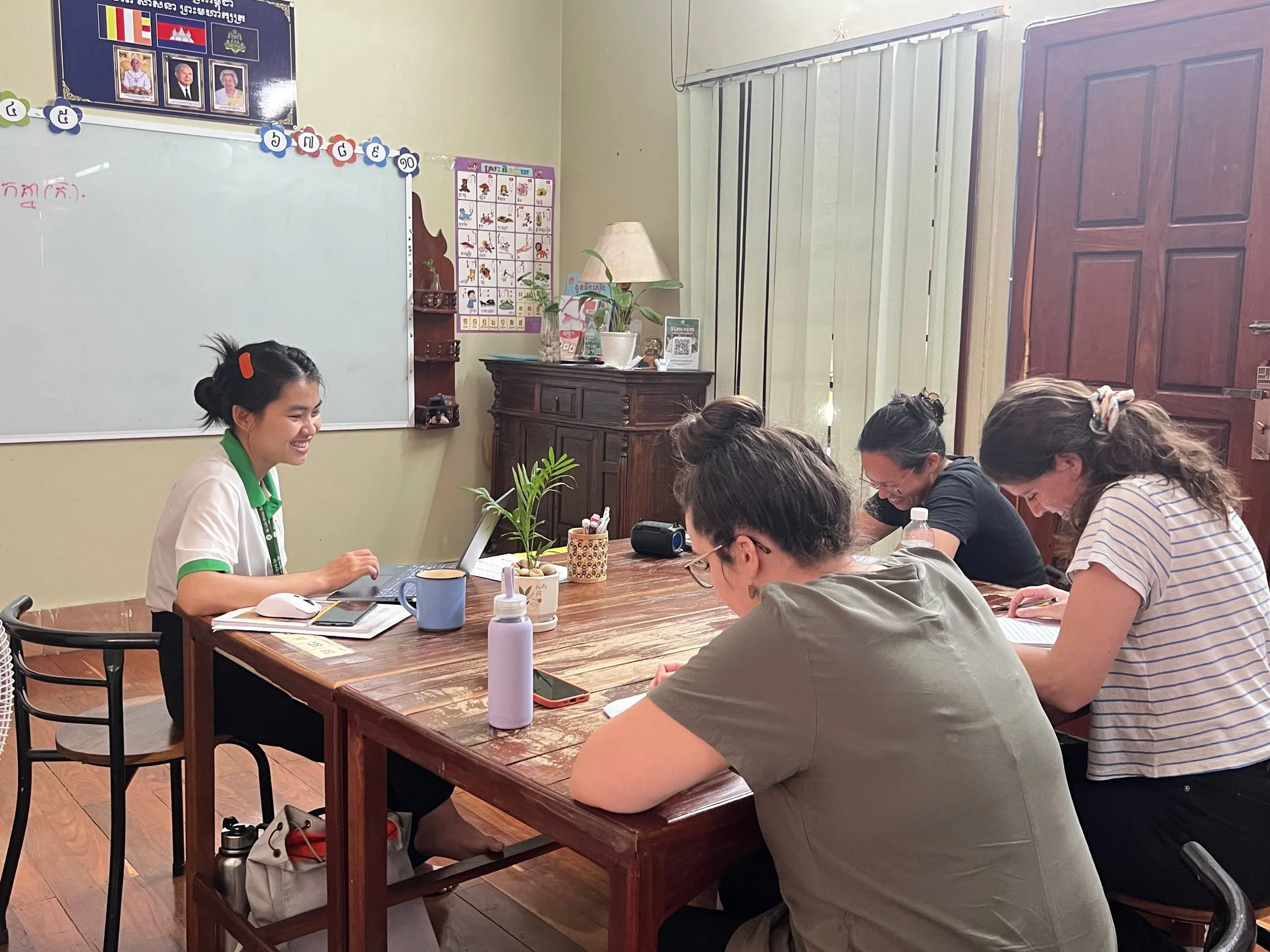 A classroom or meeting room with multiple women sitting around a rectangular wooden table, some taking notes or working on devices. One woman at the head of the table is smiling and appears to be leading the discussion. The room has a whiteboard, a decorative wooden cabinet, a potted plant, and posters on the wall. The women are engaged and focused on the activity.