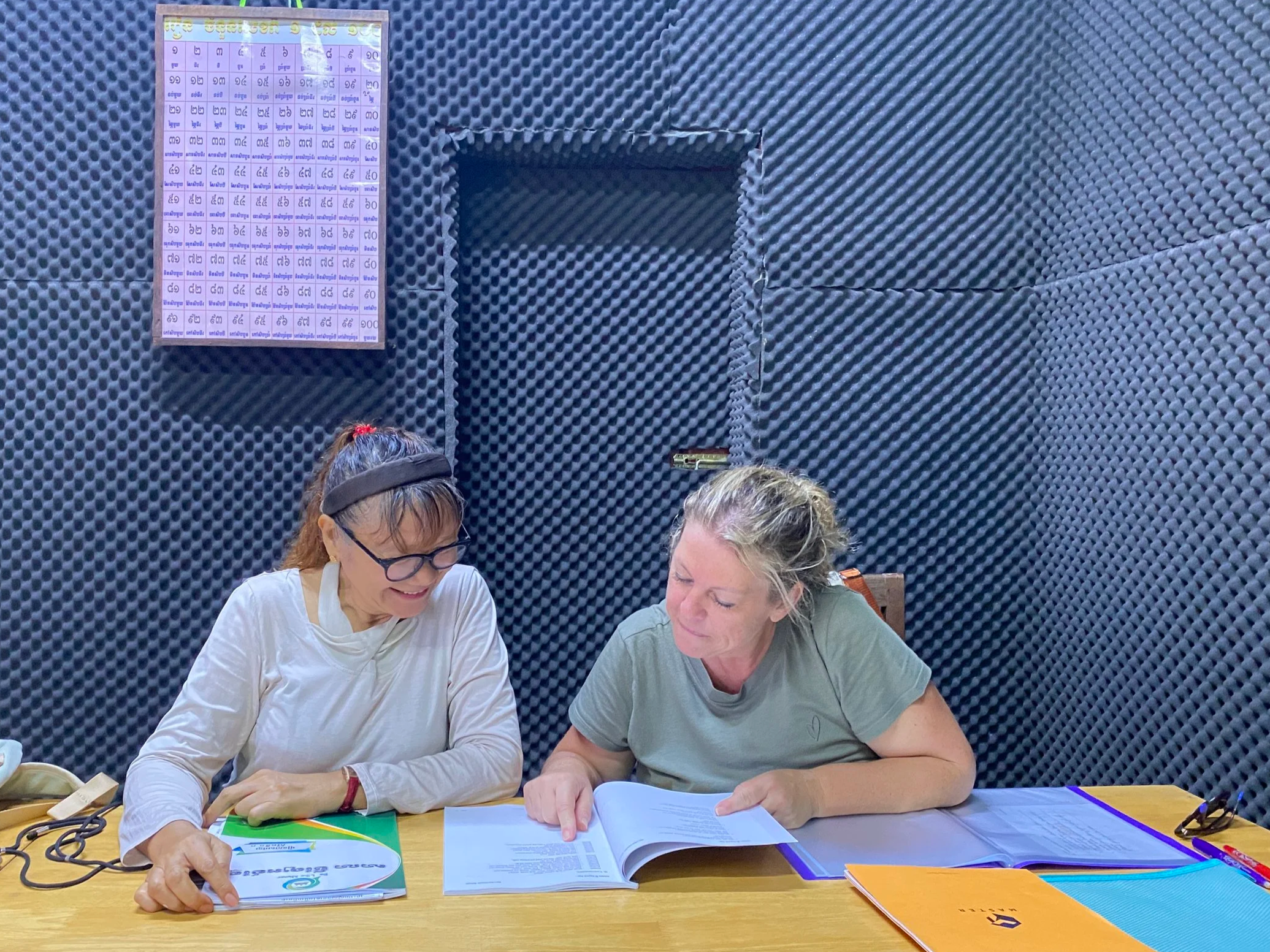 Two women sitting at a table, looking at a document together in a recording studio with soundproofing foam on the walls.