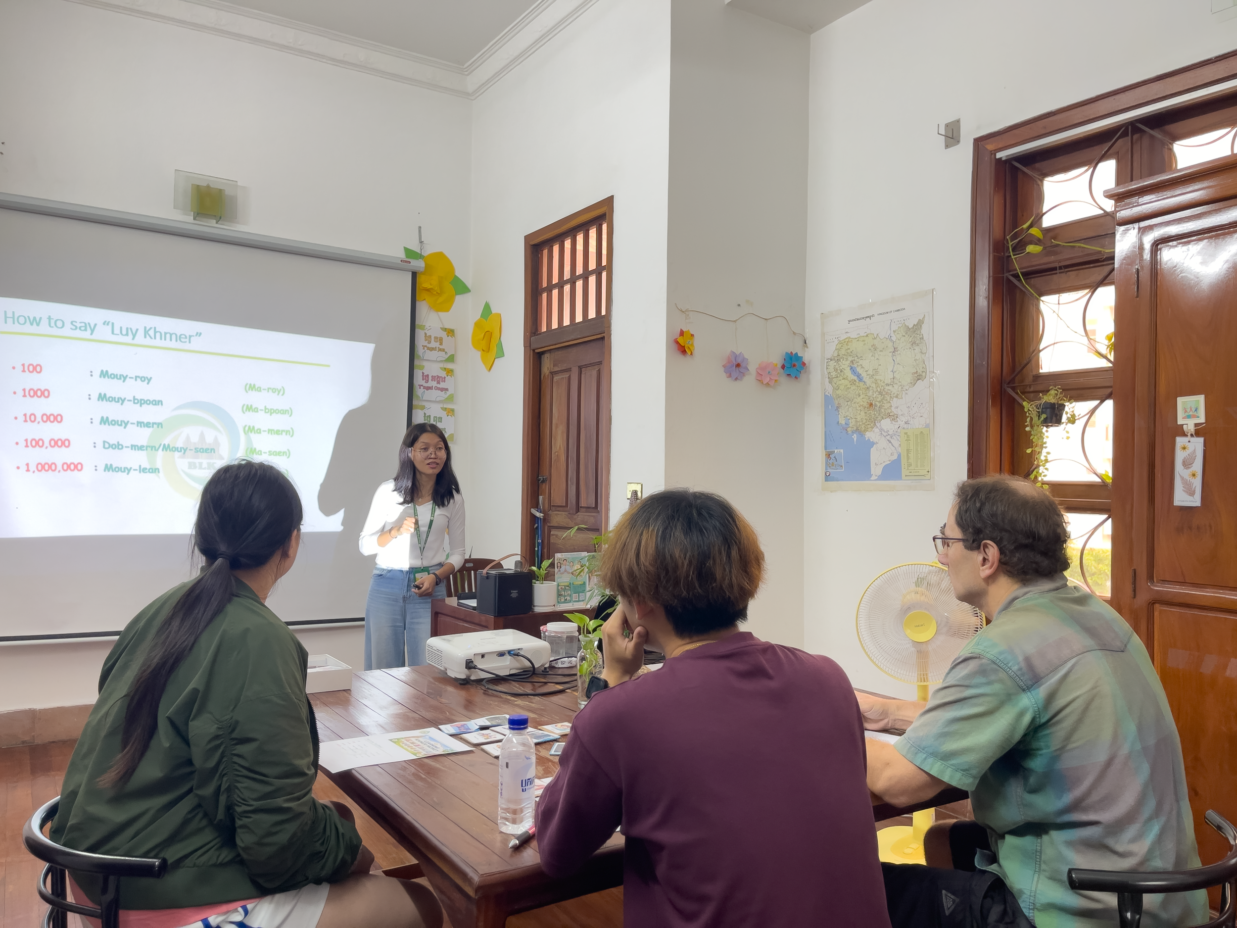 A classroom with students seated at a table, a woman standing near a projector screen giving a presentation, and a window with wooden frames and plants outside.