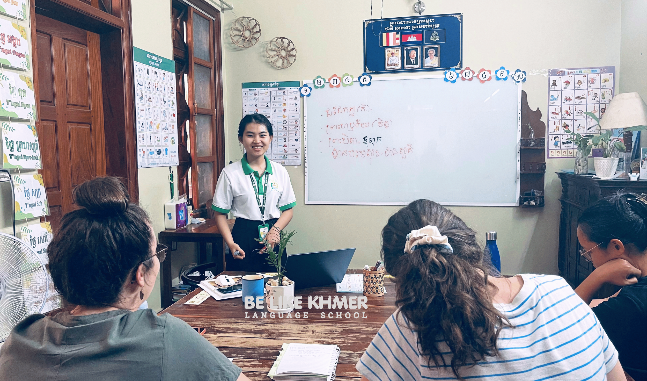 A classroom with a teacher standing in front of a whiteboard, leading a lesson for seated adult students. The room has educational posters on the walls, a wooden door, and a plant on a cabinet. The students are taking notes and listening attentively.