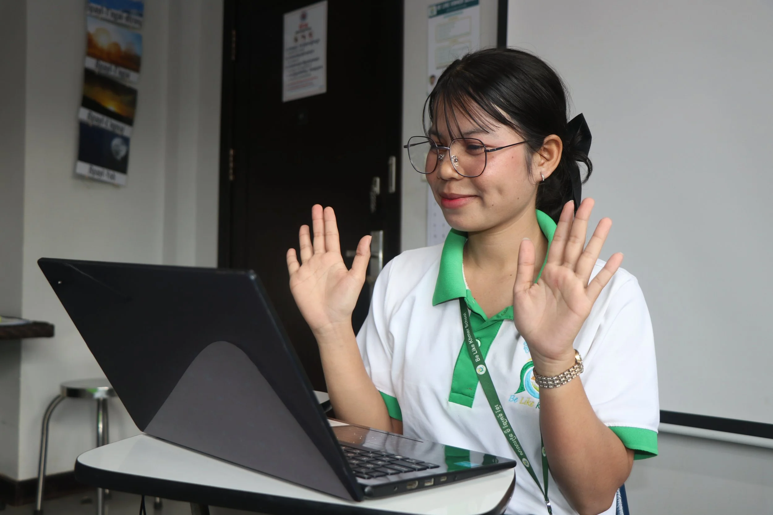 A woman with glasses, wearing a white shirt with green accents and a lanyard, is smiling and raising her hands. She is sitting at a table with a laptop in front of her. The background shows a black door, some posters on the wall, and a white wall.
