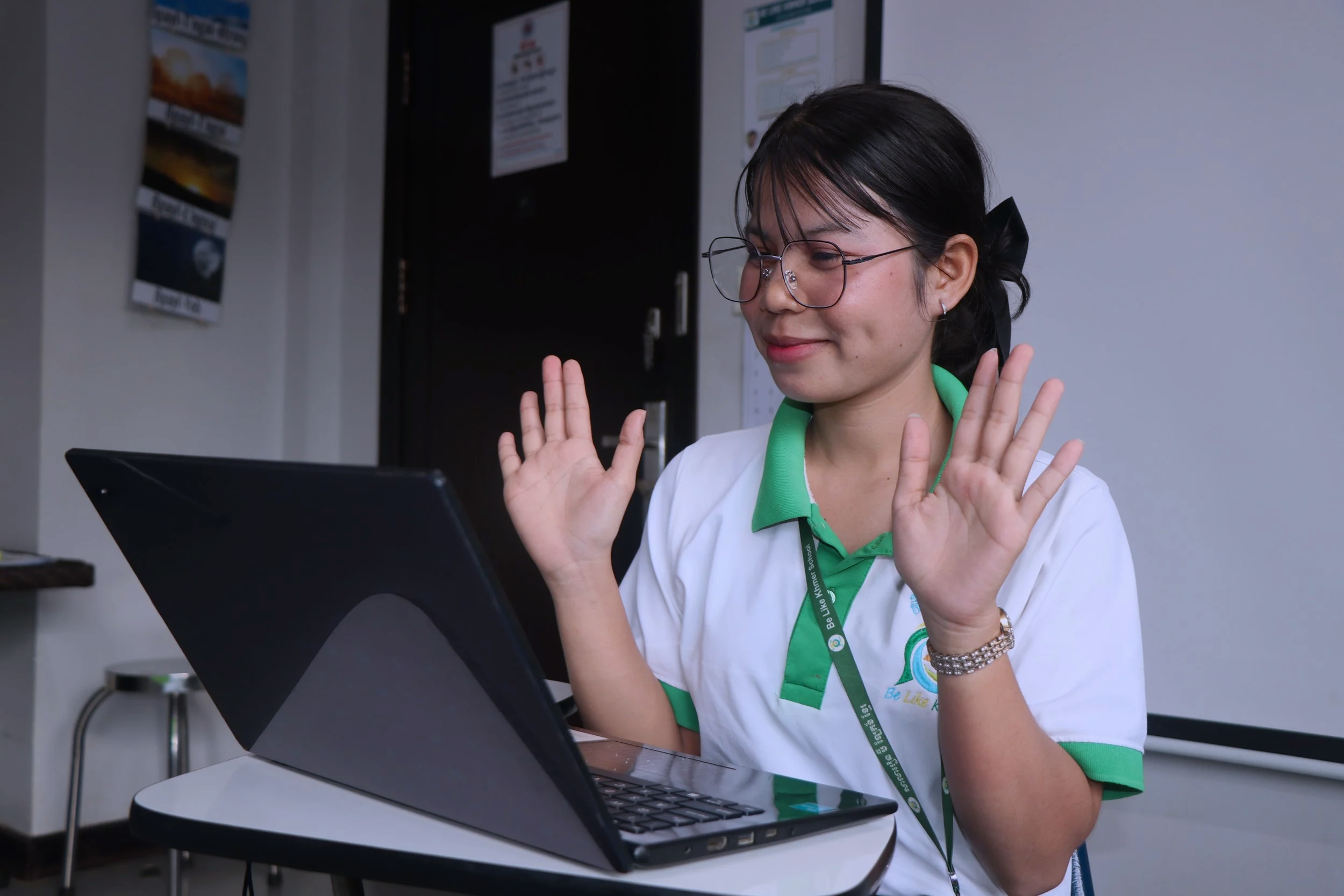 A man wearing a white polo shirt with green accents and a lanyard, sitting at a desk, smiling and gesturing with his hands while looking at his laptop with stickers on the lid. The background has a black door with papers and a white wall.