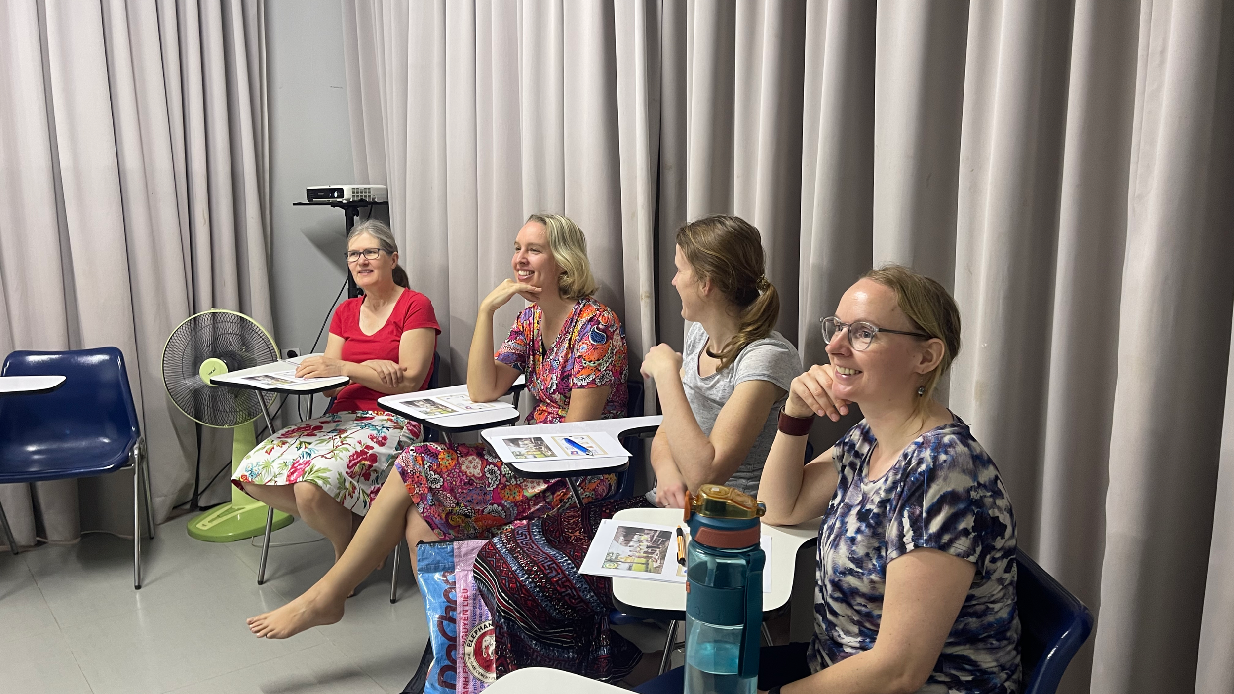 Four women sitting in a row in a meeting room with gray curtains, laughing and talking. They have papers and water bottles on their laps and tables. A fan and a projector are in the background.