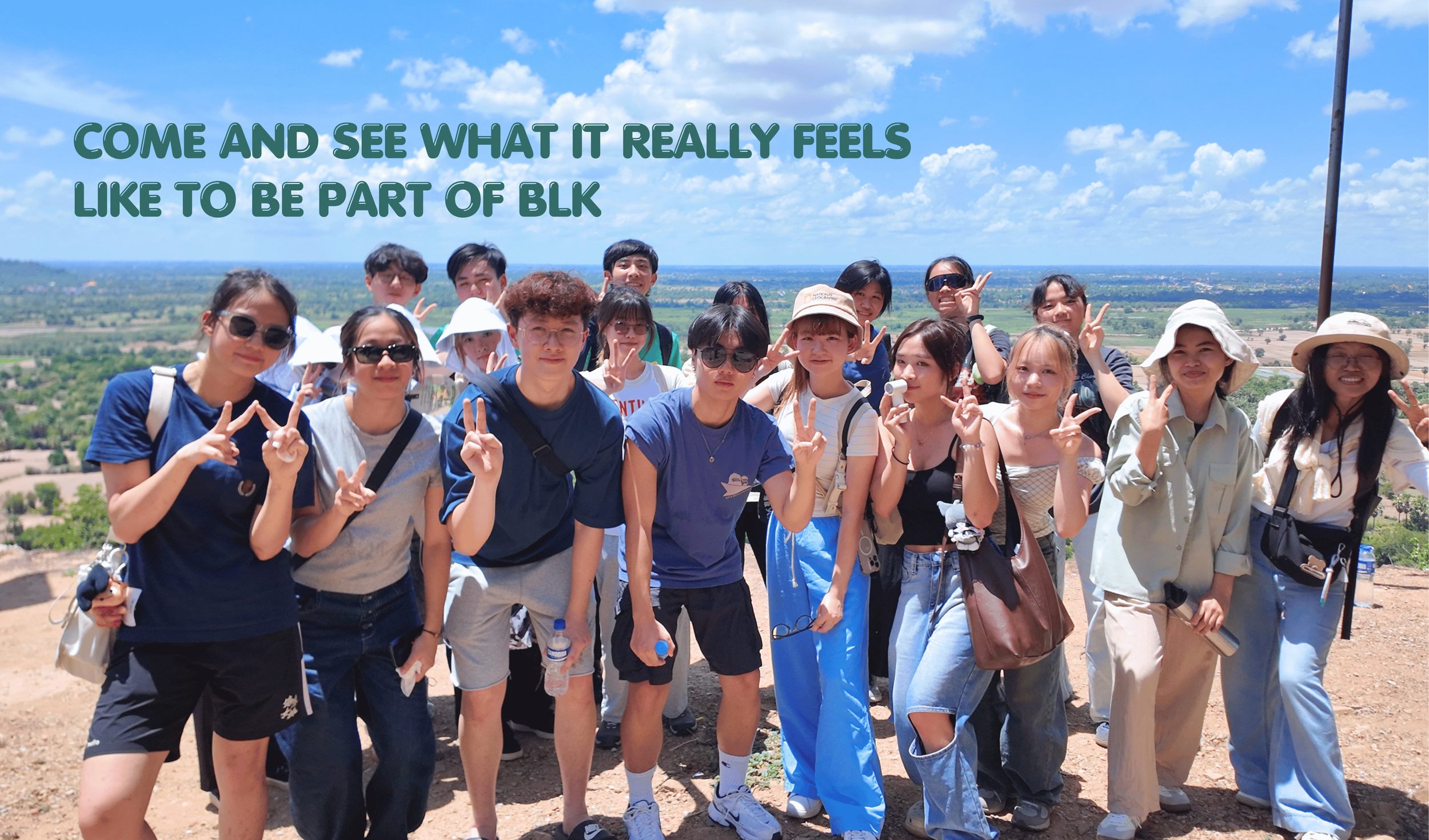 Group of young people standing outdoors on a sunny day, some making peace signs, with a vast landscape and blue sky with clouds in the background.