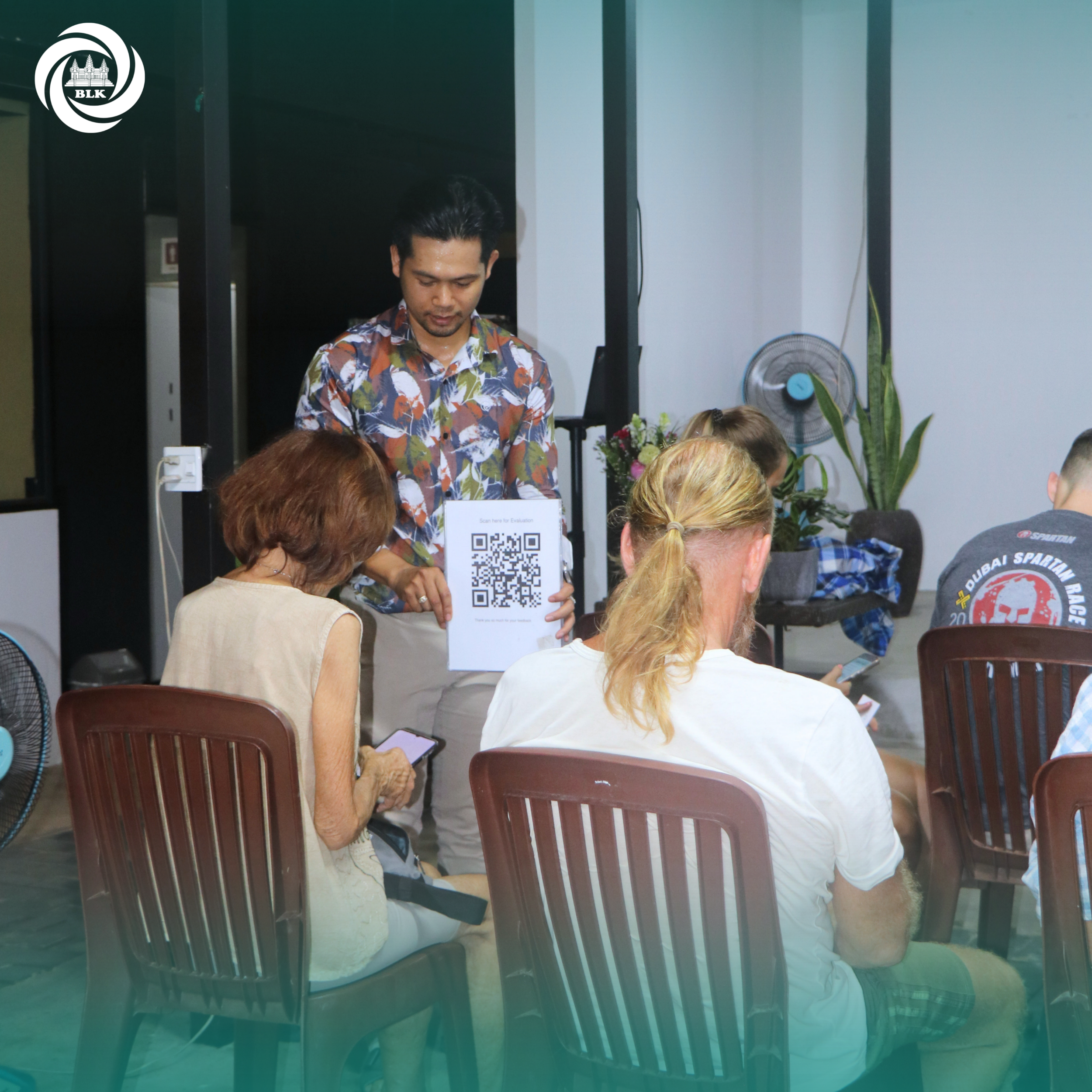 A group of people seated around tables in a room, with a man standing in the background holding a sign featuring a QR code. The logo of BLK is in the top left corner.