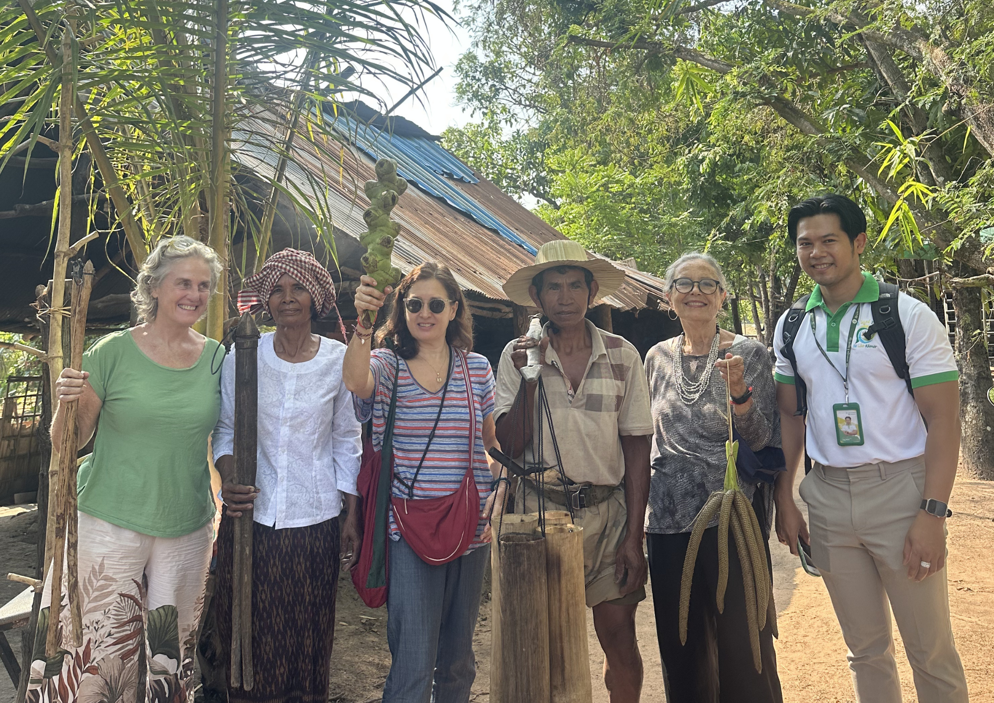 Group of six people, including women and men, standing outdoors in a rural setting with trees and a rustic house behind them. One woman holds a bunch of green bananas, and others are smiling at the camera.