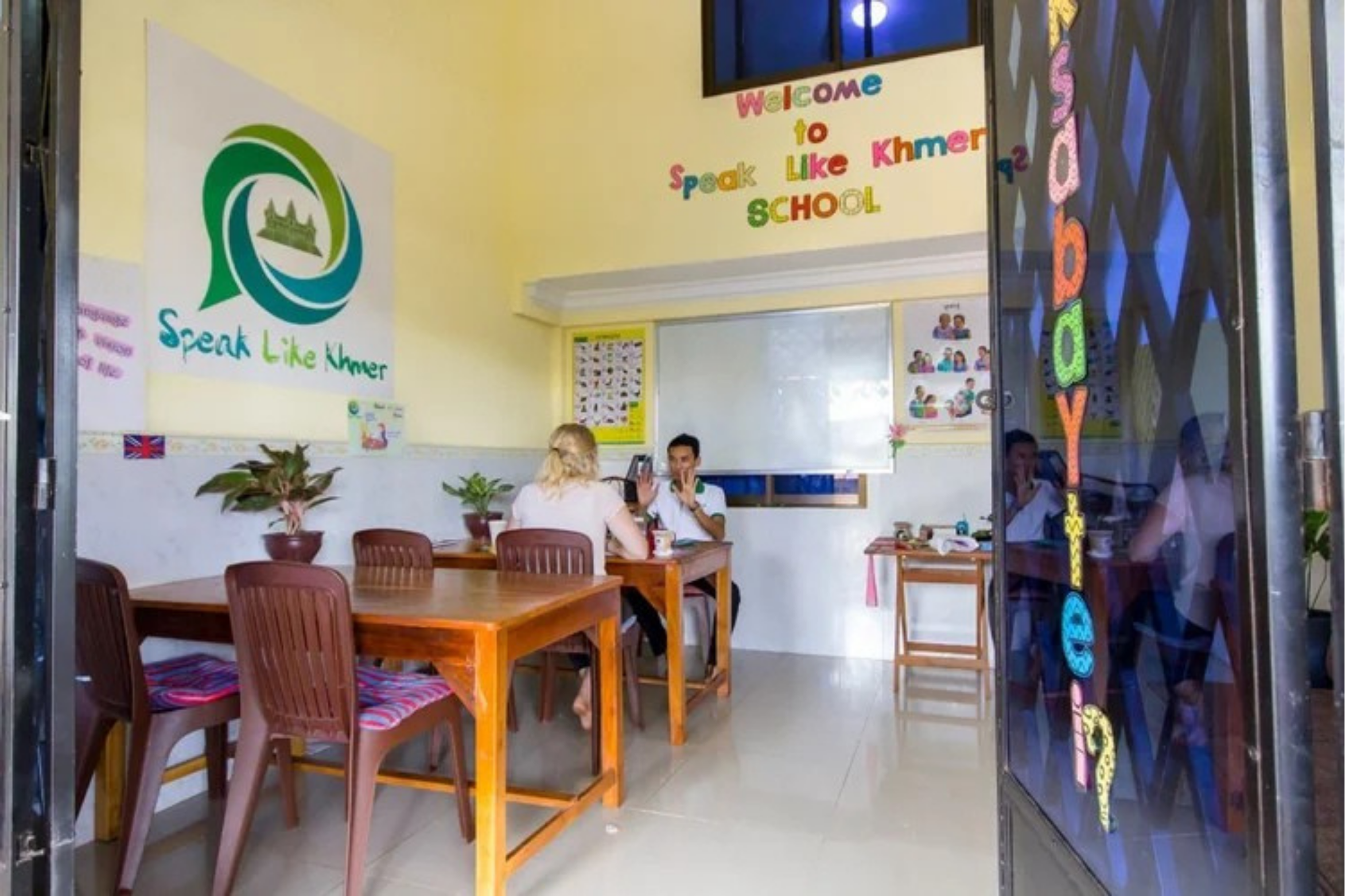Inside a classroom or office with yellow walls, showing a sign that reads 'Speak Like Khmer School' and a logo with a green icon including a castle. Two women are seated at a table, engaging in conversation, with signs and posters on the wall.