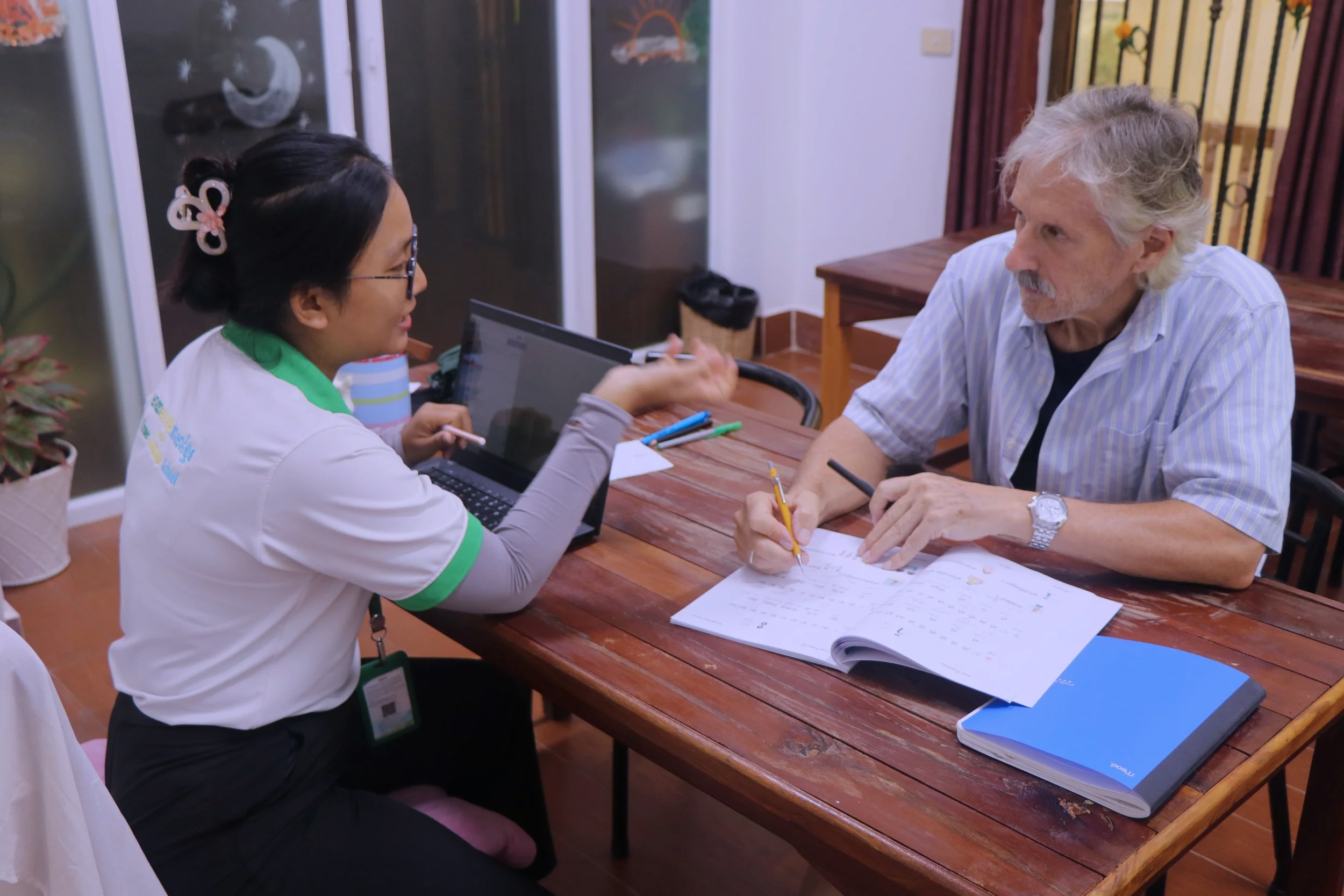 A woman and an older man sitting at a wooden table, engaged in conversation. The woman is smiling, holding a pen, and wearing glasses and a white shirt with green accents. The man has gray hair and a beard, wearing a striped shirt, and holding a yellow pencil. There are papers, notebooks, and writing instruments on the table, and a laptop partially visible. The background shows a glass cabinet and wooden railing.