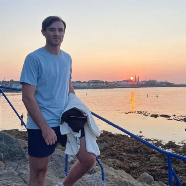 Young man standing on rocks by the water at sunset, holding a jacket, with city skyline in the background.