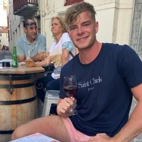 A young man sitting outdoors at a table with three older adults, holding a glass of red wine, smiling at the camera.