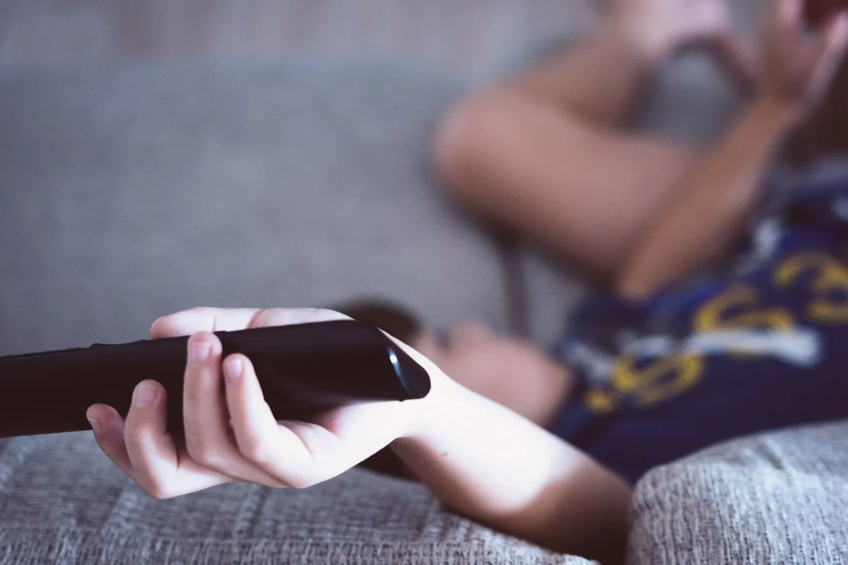 Close-up of a person holding a TV remote control while reclining on a sofa, with the background intentionally blurred.