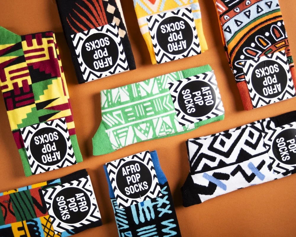 Multiple pairs of colorful socks with African-inspired patterns displayed on an orange background, each pair labeled 'AFRO POP SOCKS'.