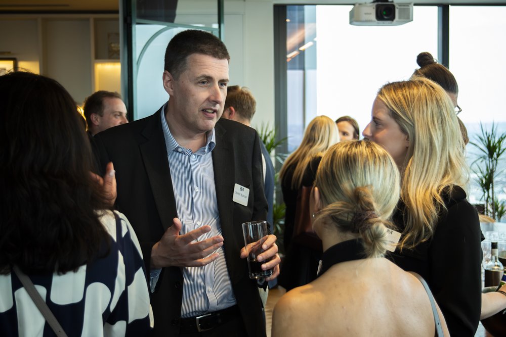 A man in a black suit and striped shirt talking to a group of women at a networking event or conference, with large windows and plants in the background.