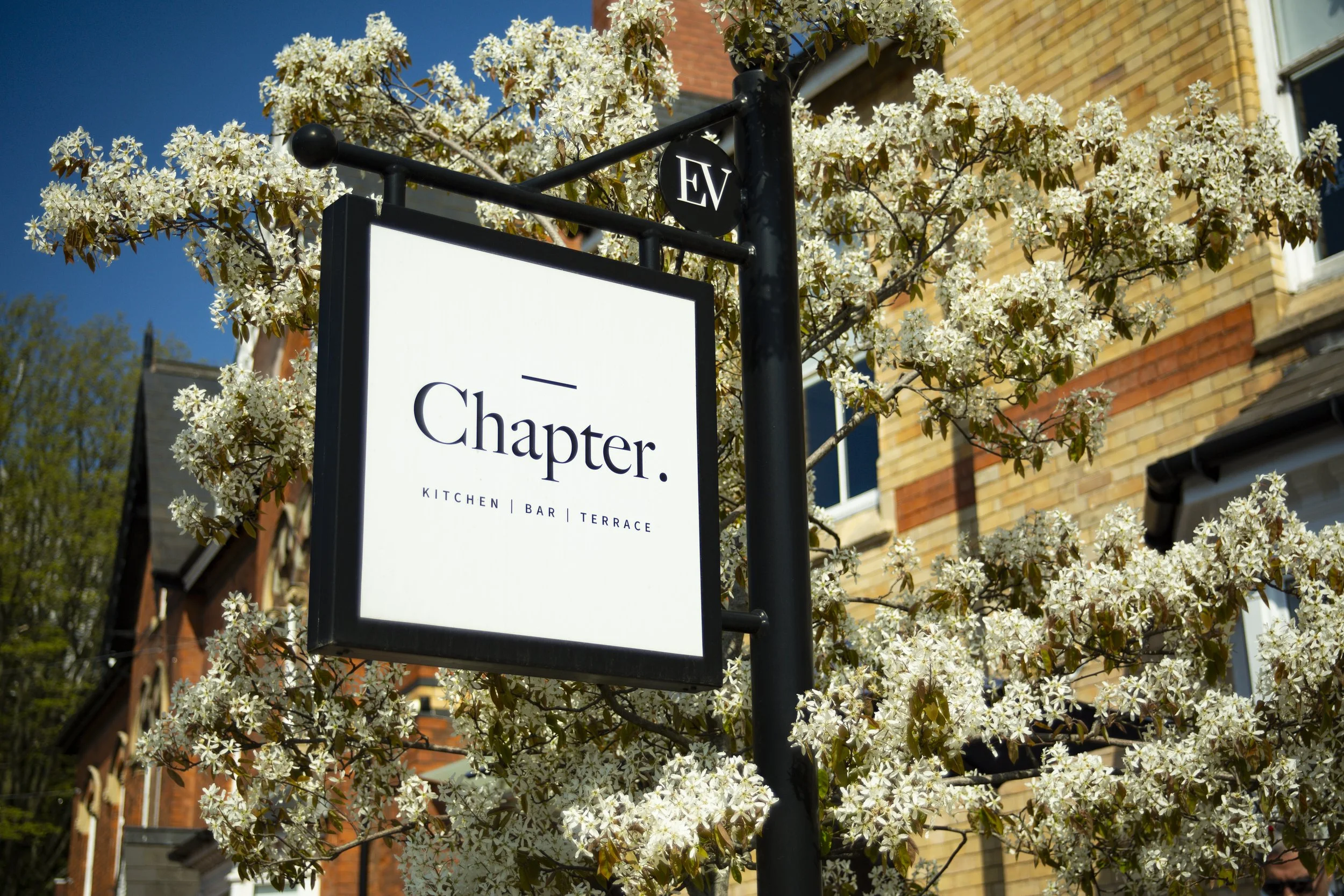 A sign for the restaurant Chapter, which features a kitchen, bar, and terrace, is hanging from a black pole surrounded by white flowering tree blossoms on a sunny day.