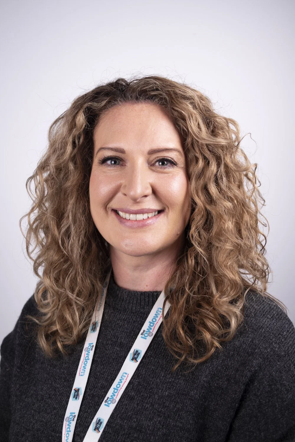 A woman with curly, light brown hair and blue eyes smiling, wearing a dark sweater and a lanyard that says 'The Show'.