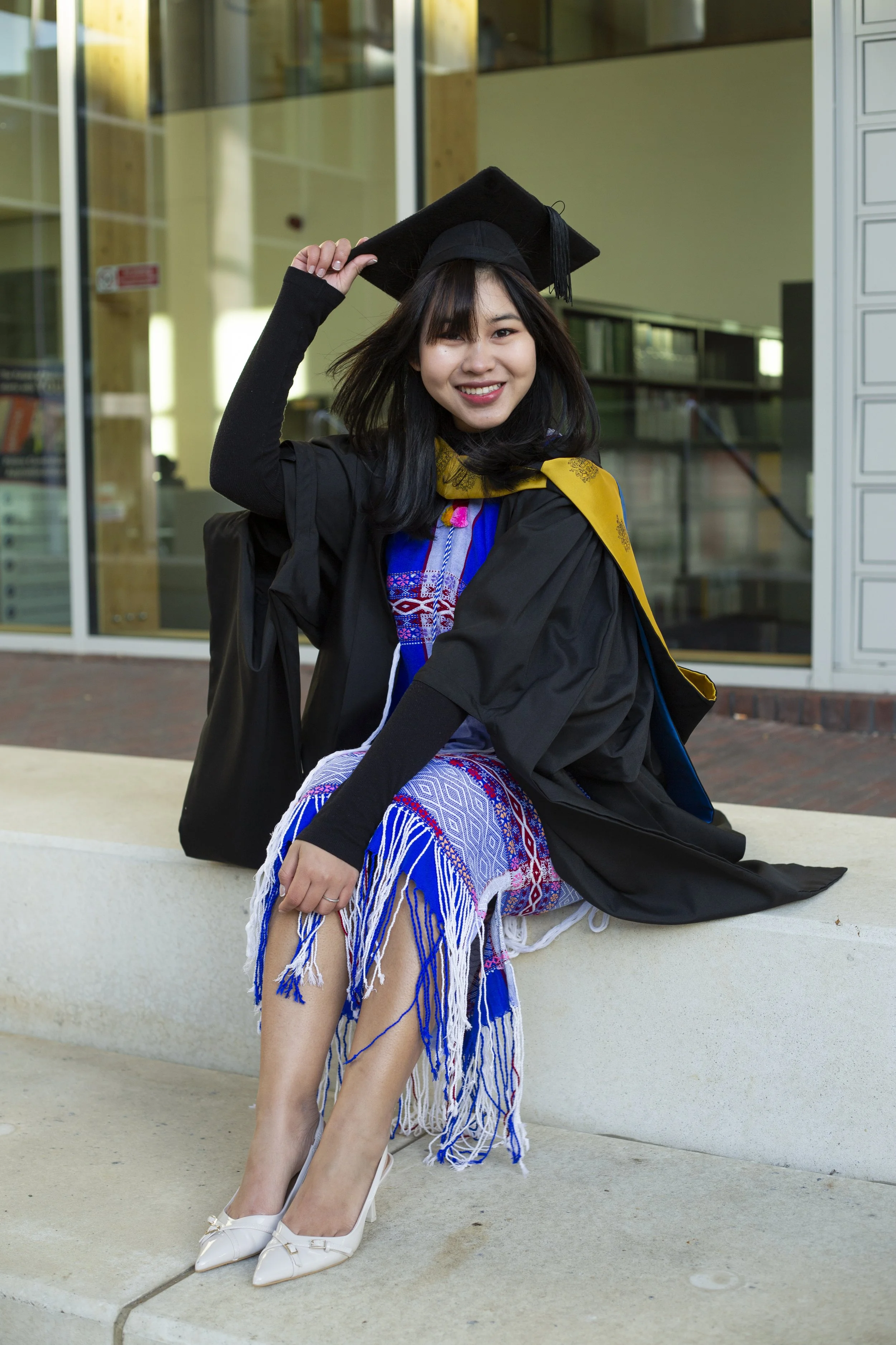 Young woman in graduation gown and cap sitting outdoors, smiling, in front of glass building.