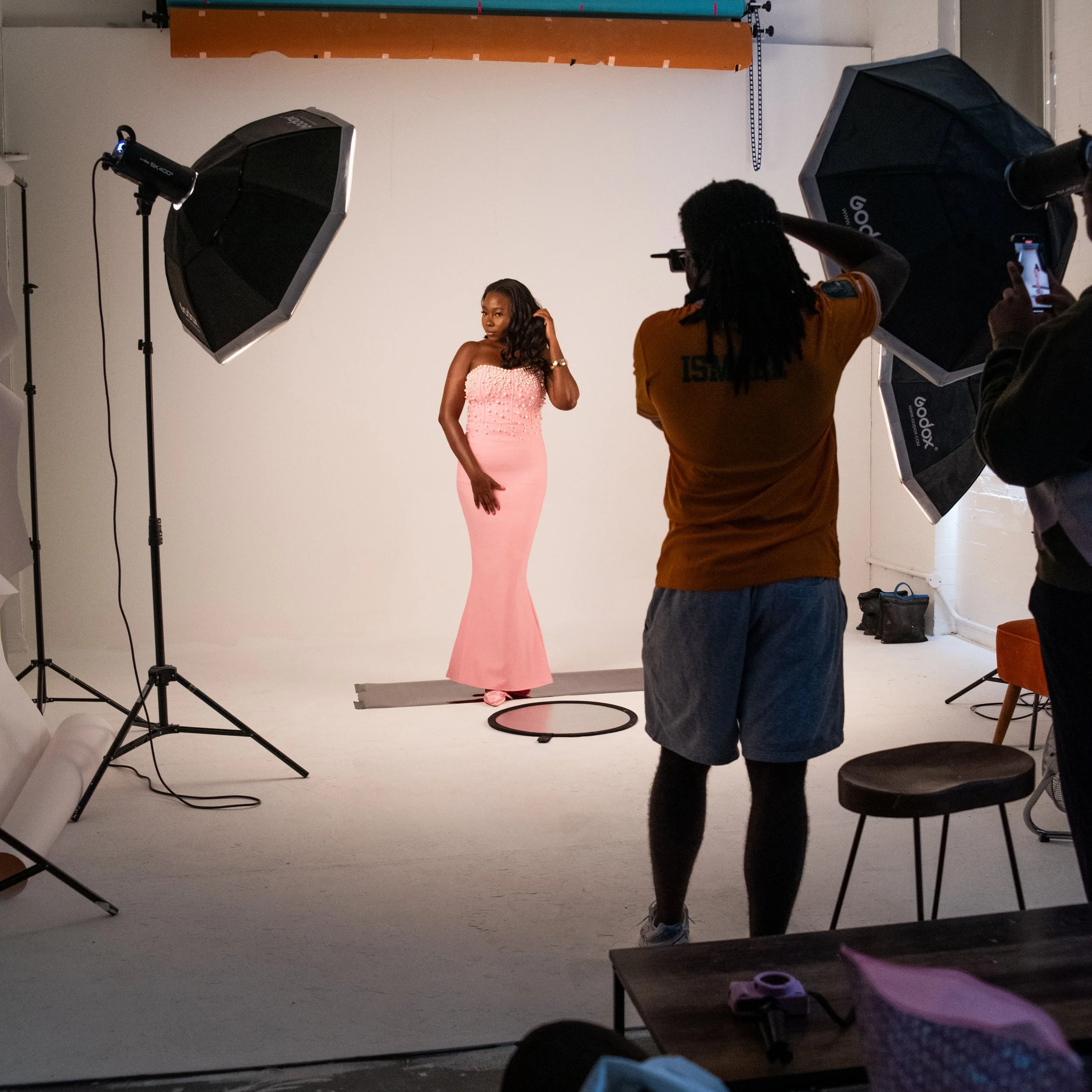 A photoshoot in progress with a woman in a pink gown posing in front of a white backdrop, with two photographers capturing her image under professional lighting setup.