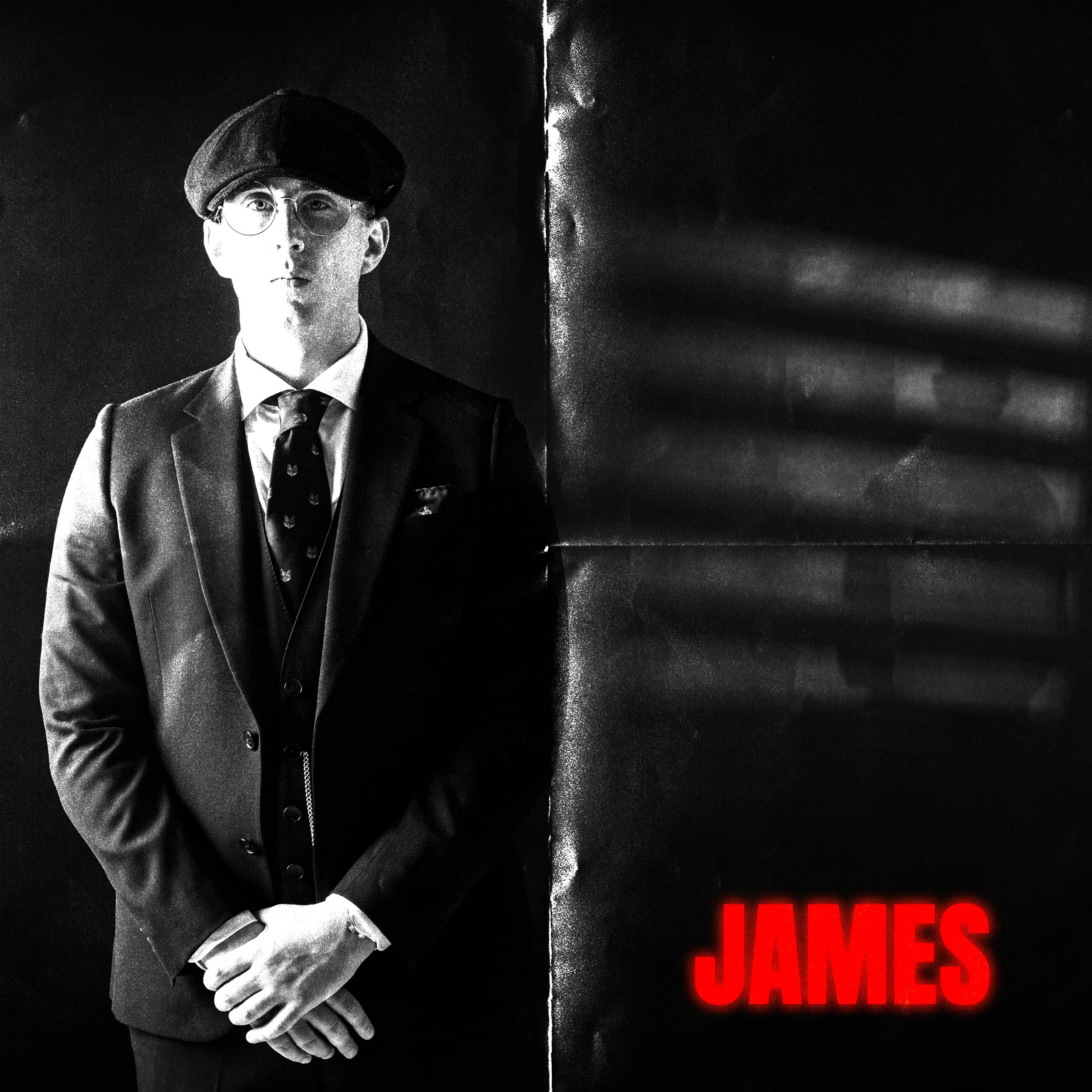 A young man in a suit, tie, and glasses wearing a beret, standing against a dark wall. The word "JAMES" is illuminated in red at the bottom right corner.