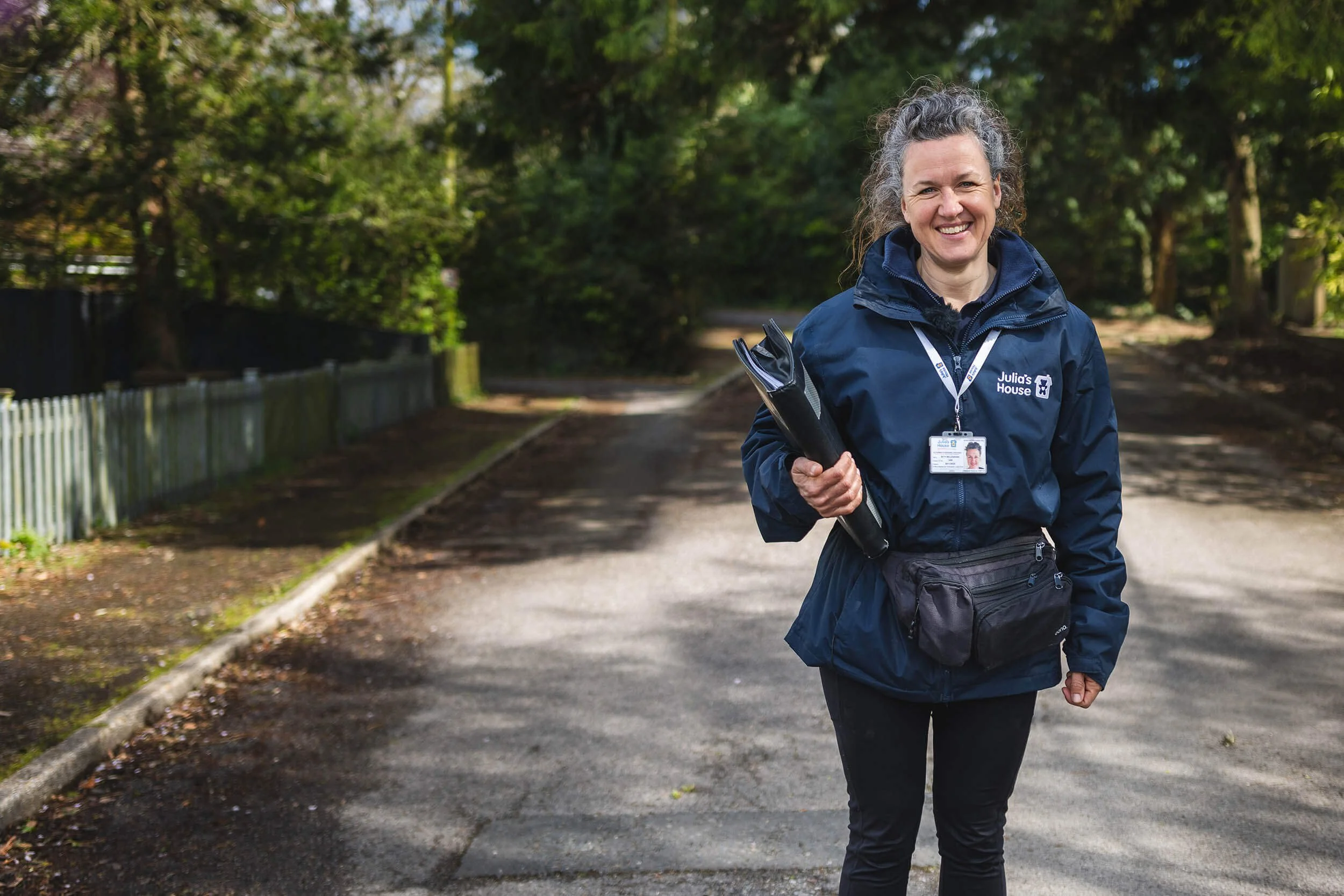 A female LFS face-to-face fundraiser, smiling, wearing a navy jacket with a badge and a pouch around her waist, standing on a paved path in a wooded area, holding a binder.