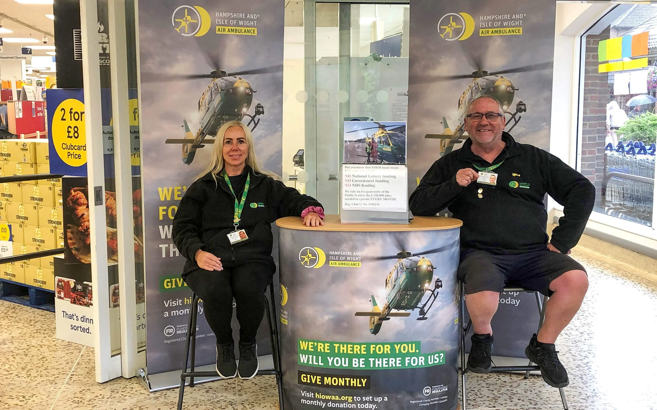 Two representatives from Hampshire and Isle of Wight Air Ambulance sitting at a promotional booth inside a store, with large banners featuring a helicopter above and charity information displayed.