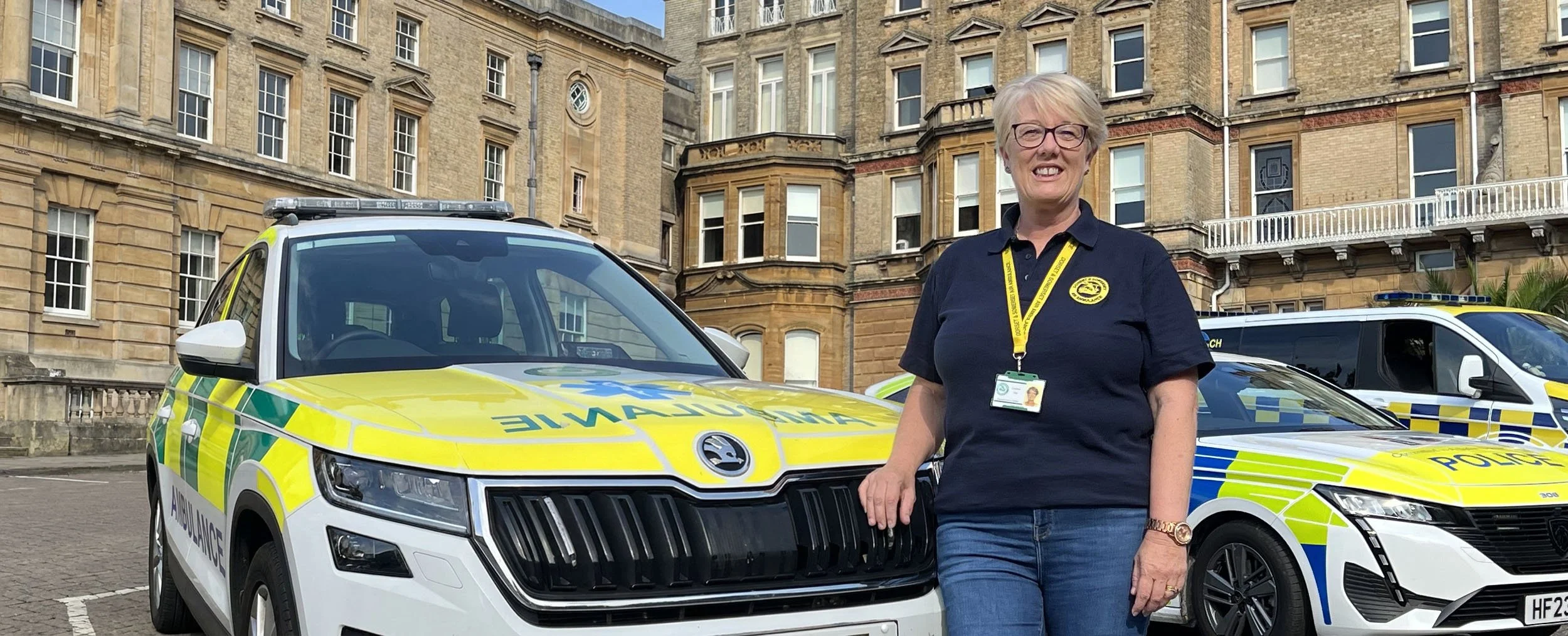 Caroline Guy standing outdoors in front of emergency response vehicles, including an ambulance and a police car, in front of a historic brick building.